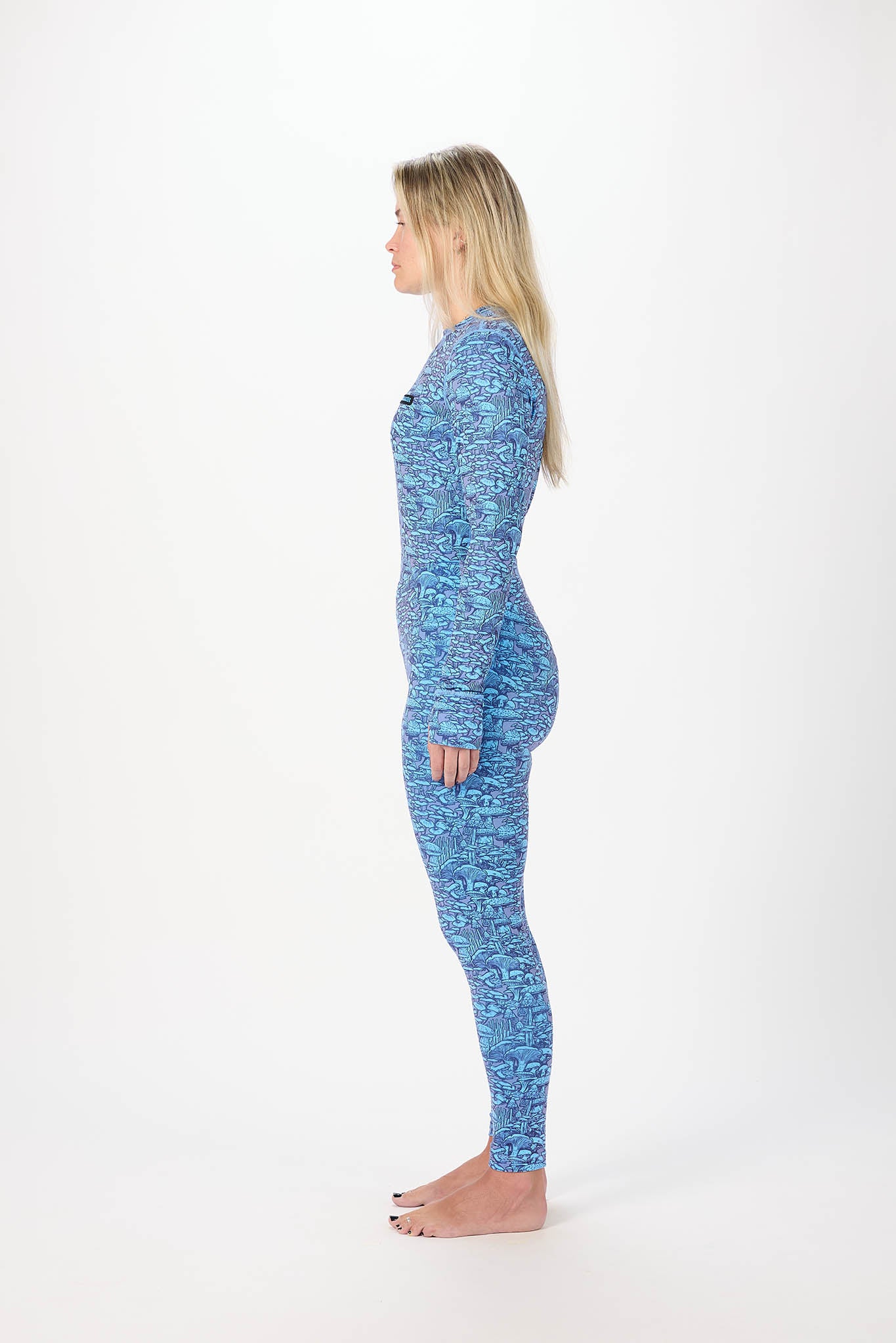 A woman stands in profile against a plain white studio background, wearing a light blue long-sleeve top with a colorful fruit print and mushroom leggings.