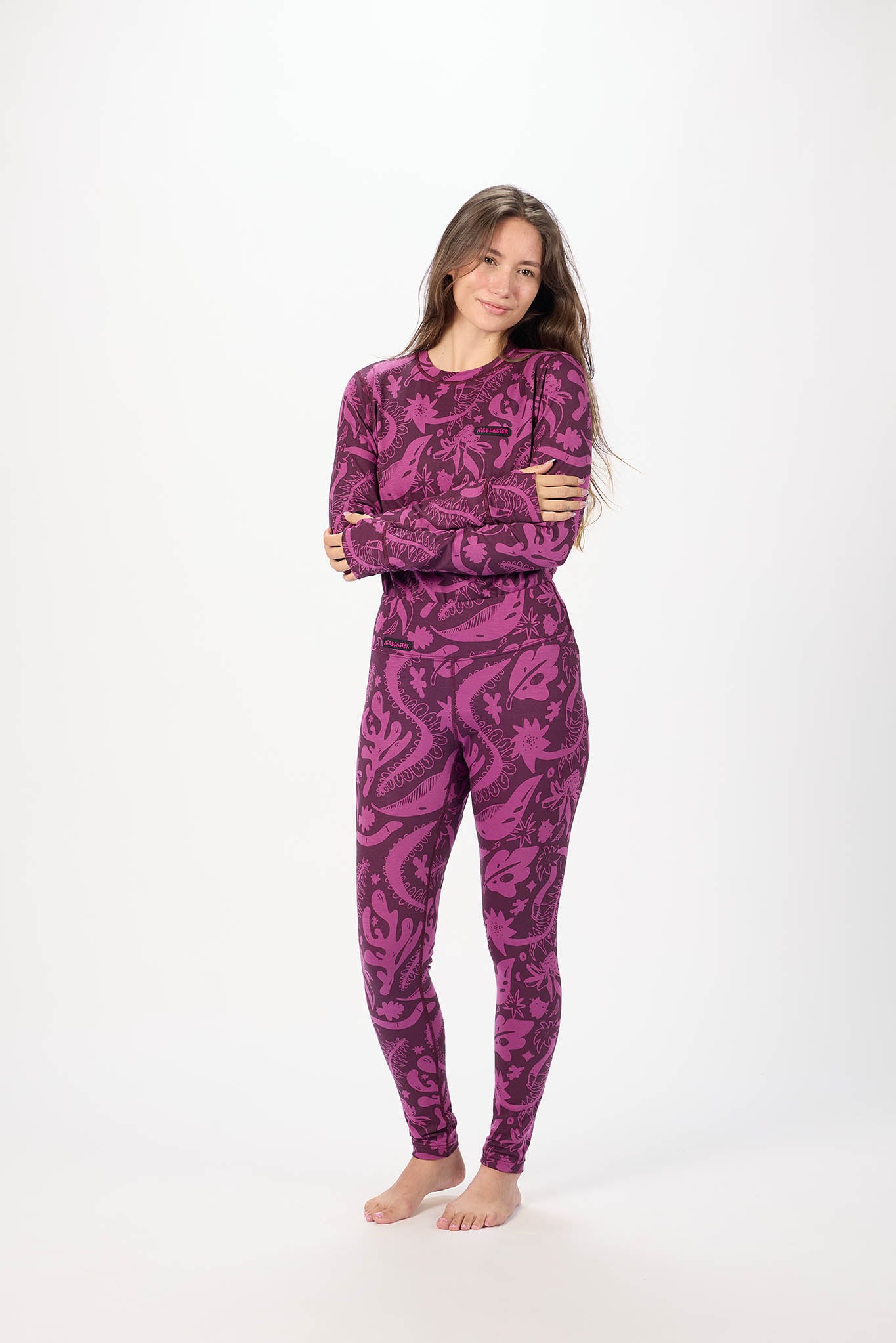 A woman modeling a matching two-piece base Layer set with a purple and magenta paisley pattern, standing against a plain white studio background.
