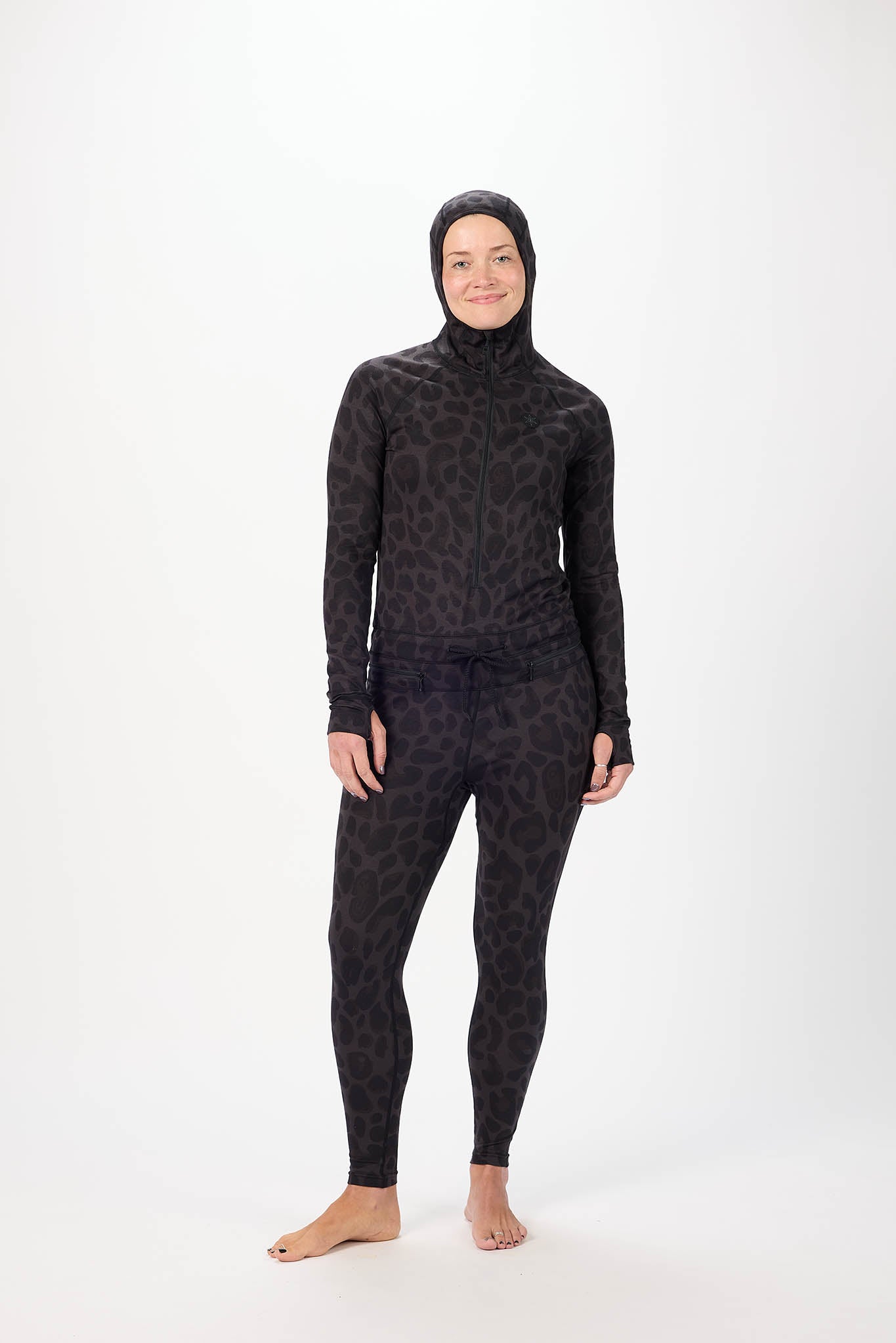A woman wearing a full-body black leopard-print onesie with a hood and face mask, standing against a plain white studio background.
