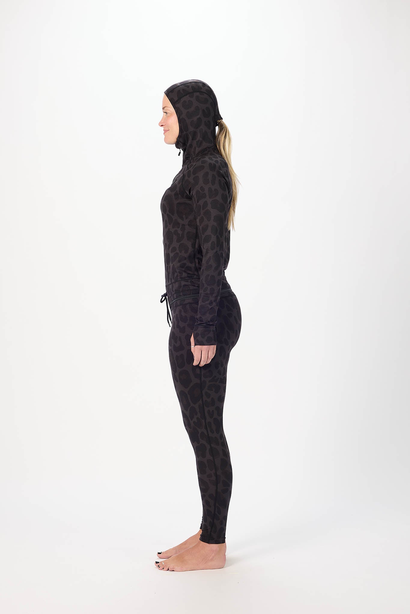 A woman in side wearing a full-body black leopard-print onesie with a hood and face mask, standing against a plain white studio background.