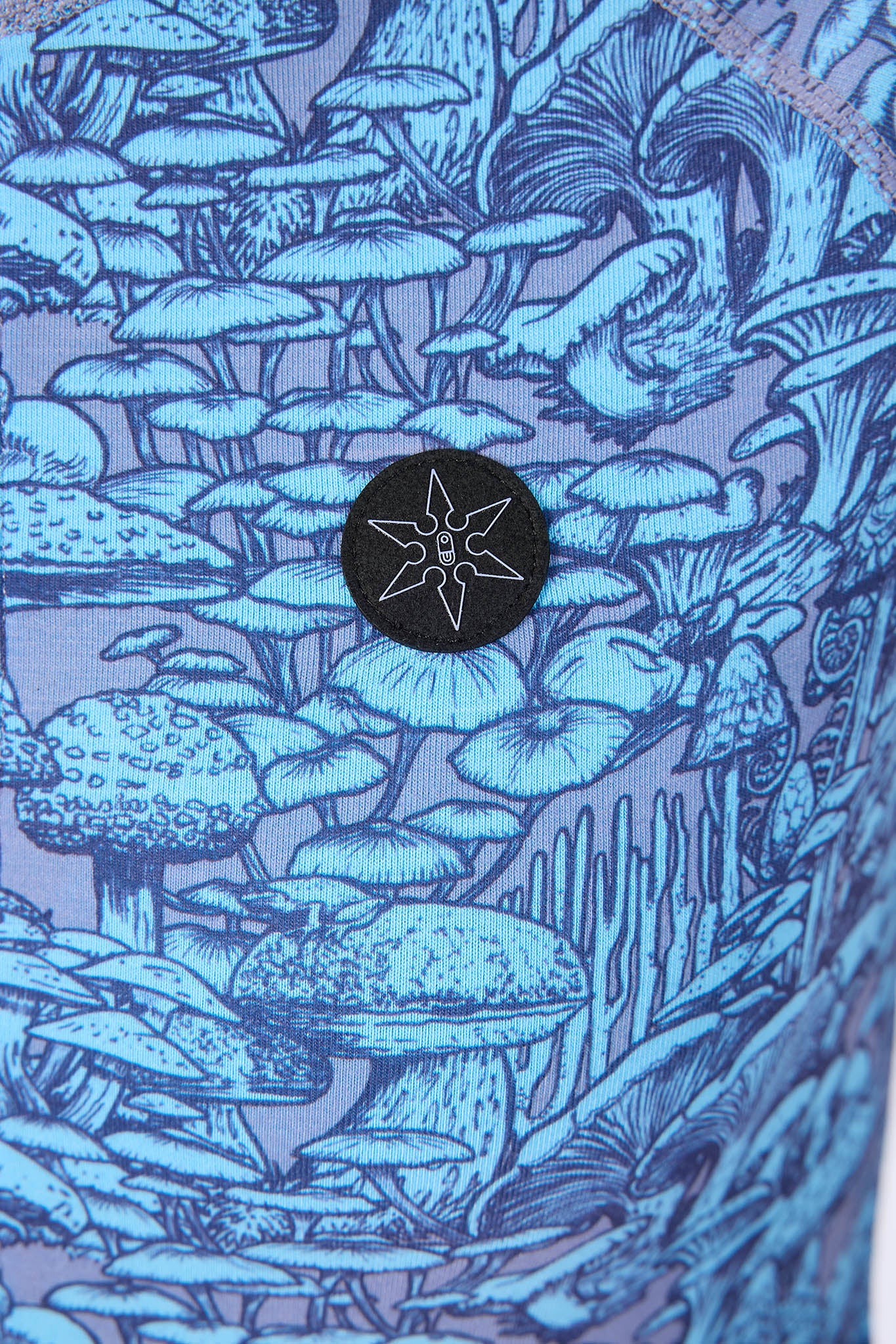 Close-up of a blue and purple patterned fabric featuring an illustrated mushroom forest, with a black circular patch containing a white geometric star-like logo.