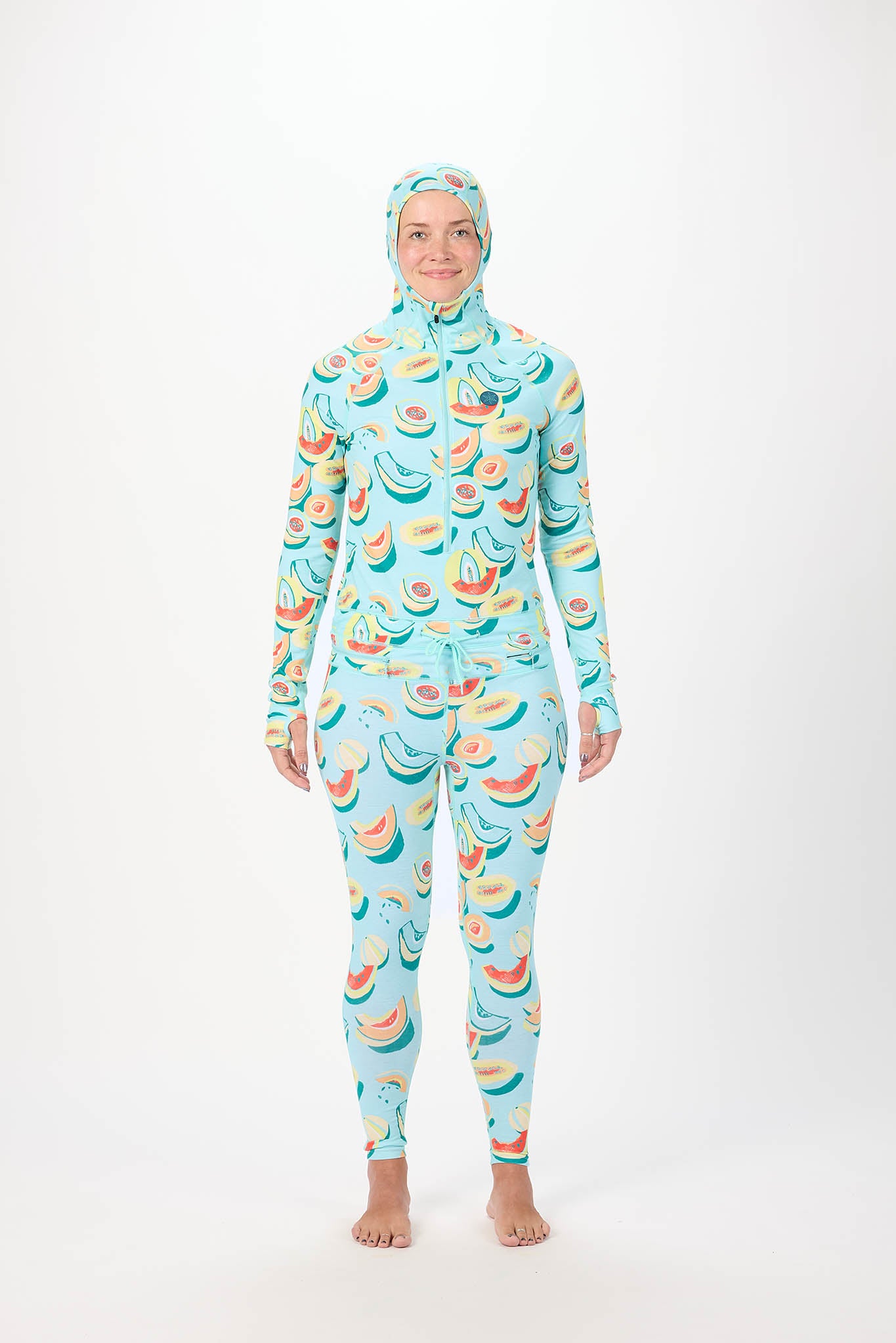 A woman modeling a full-body hooded one-piece with a light white background and a repeating print of bananas, watermelons, and strawberries, standing against a plain white studio background.