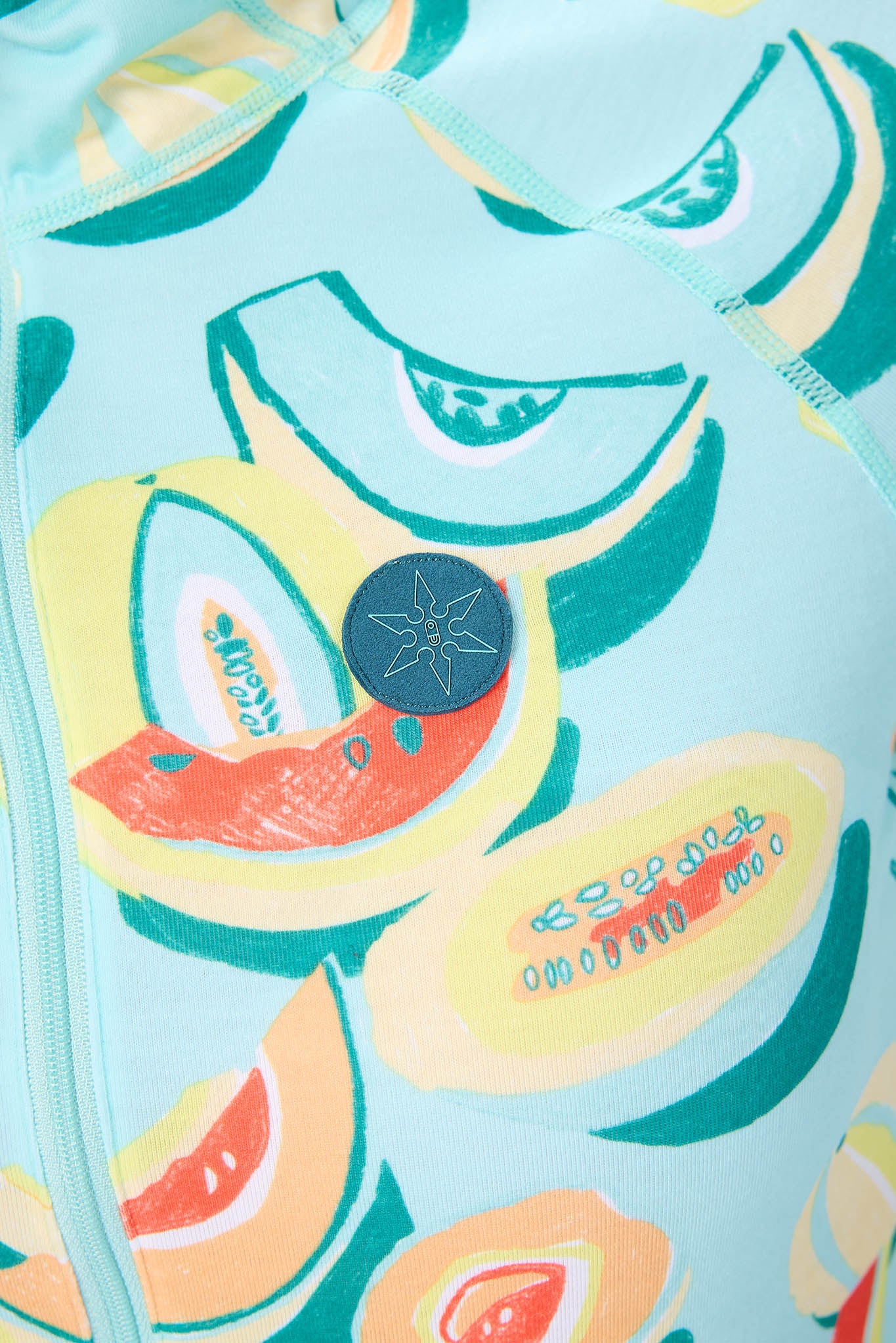 Close-up of a light aqua one-piece swimsuit featuring a colorful avocado print in shades of green, yellow, orange, and white, with a dark blue circular logo patch on the chest.