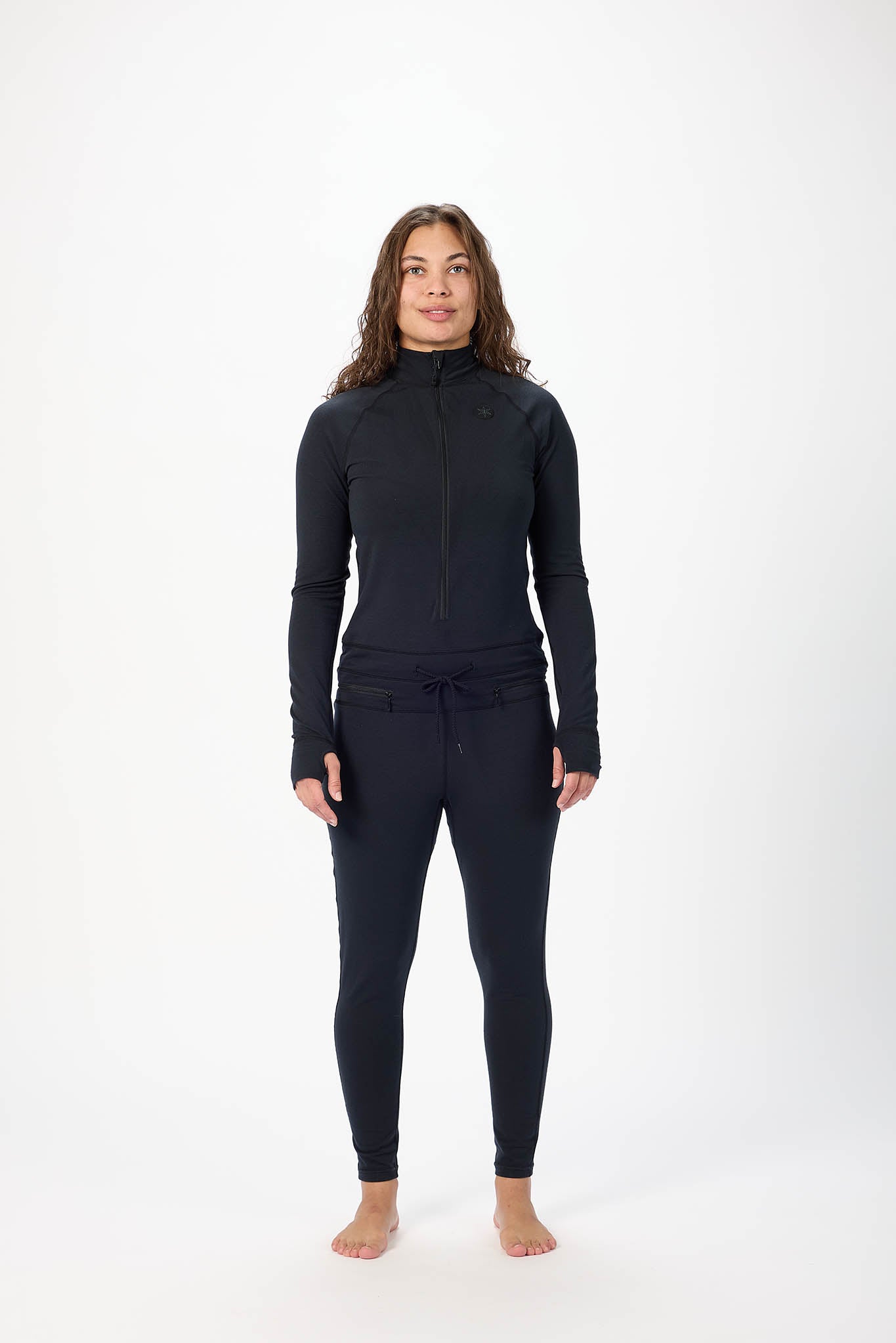 A woman stands barefoot against a plain white studio background wearing a dark navy long-sleeve zip-front athletic top and matching dark navy leggings.