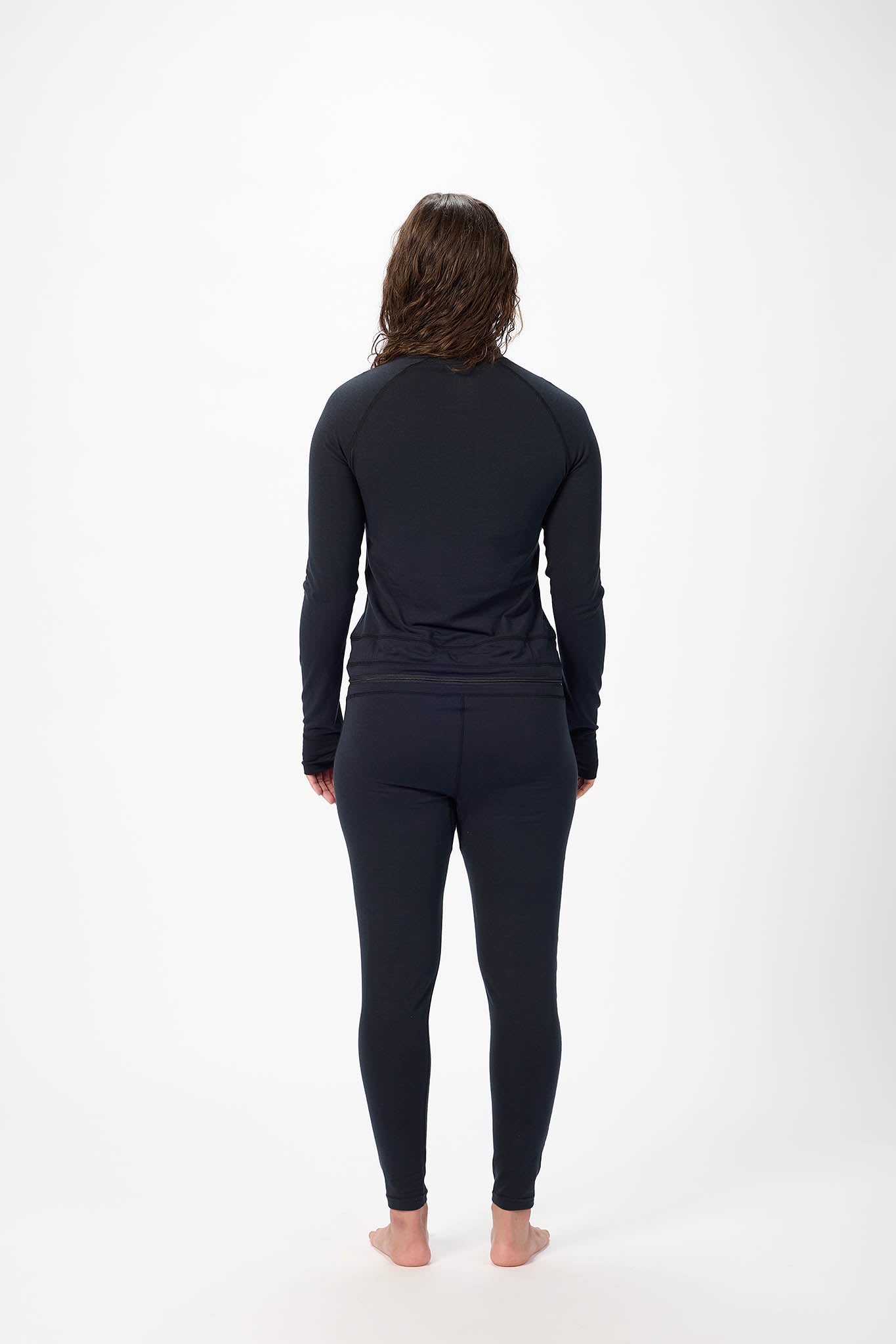 A woman viewed from behind wearing a matching black long-sleeve top and high-waisted leggings set, standing barefoot against a plain white studio background.