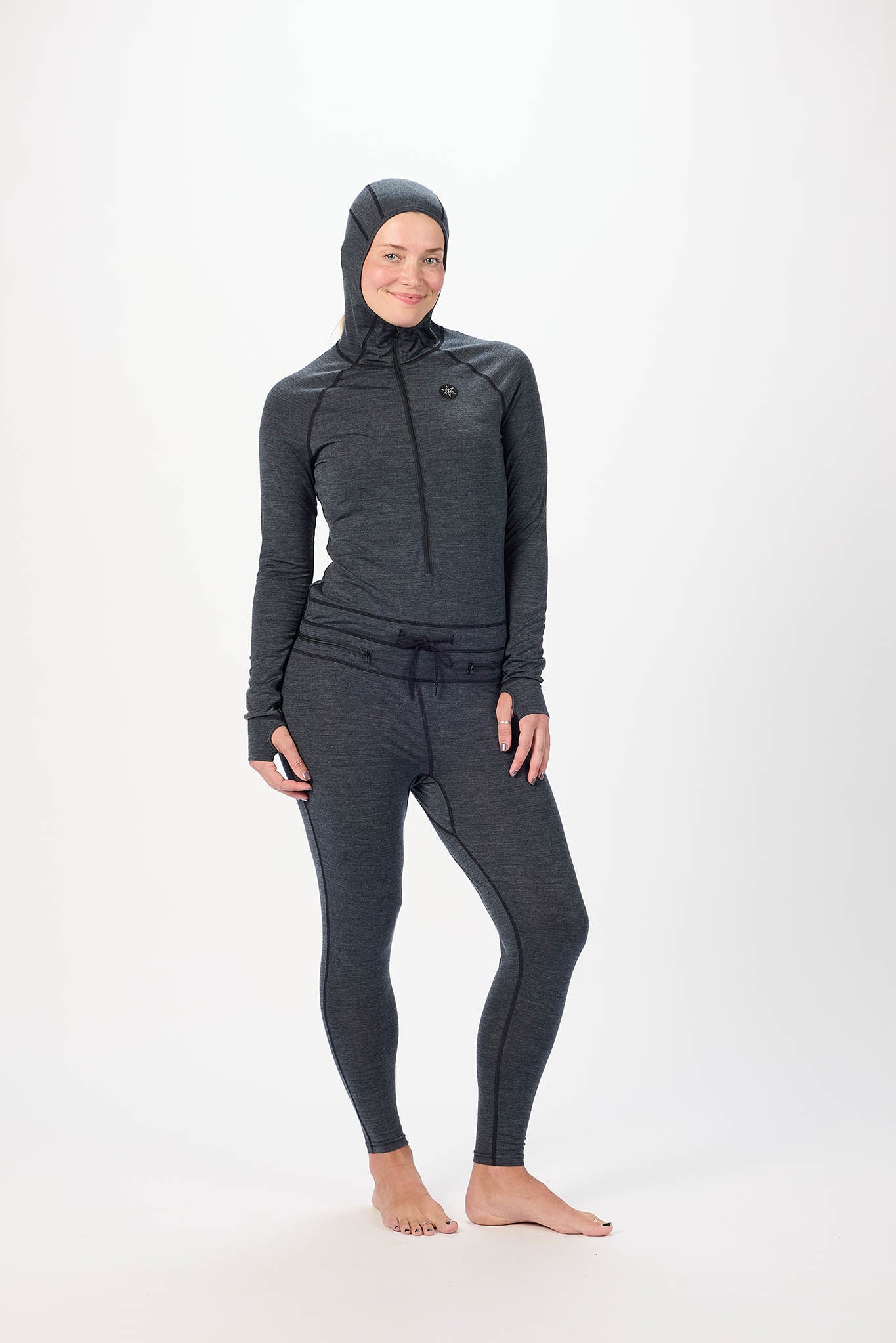 A woman modeling a dark gray full-body onesie with a hood, long sleeves, and full-length leggings, standing against a plain white studio background.