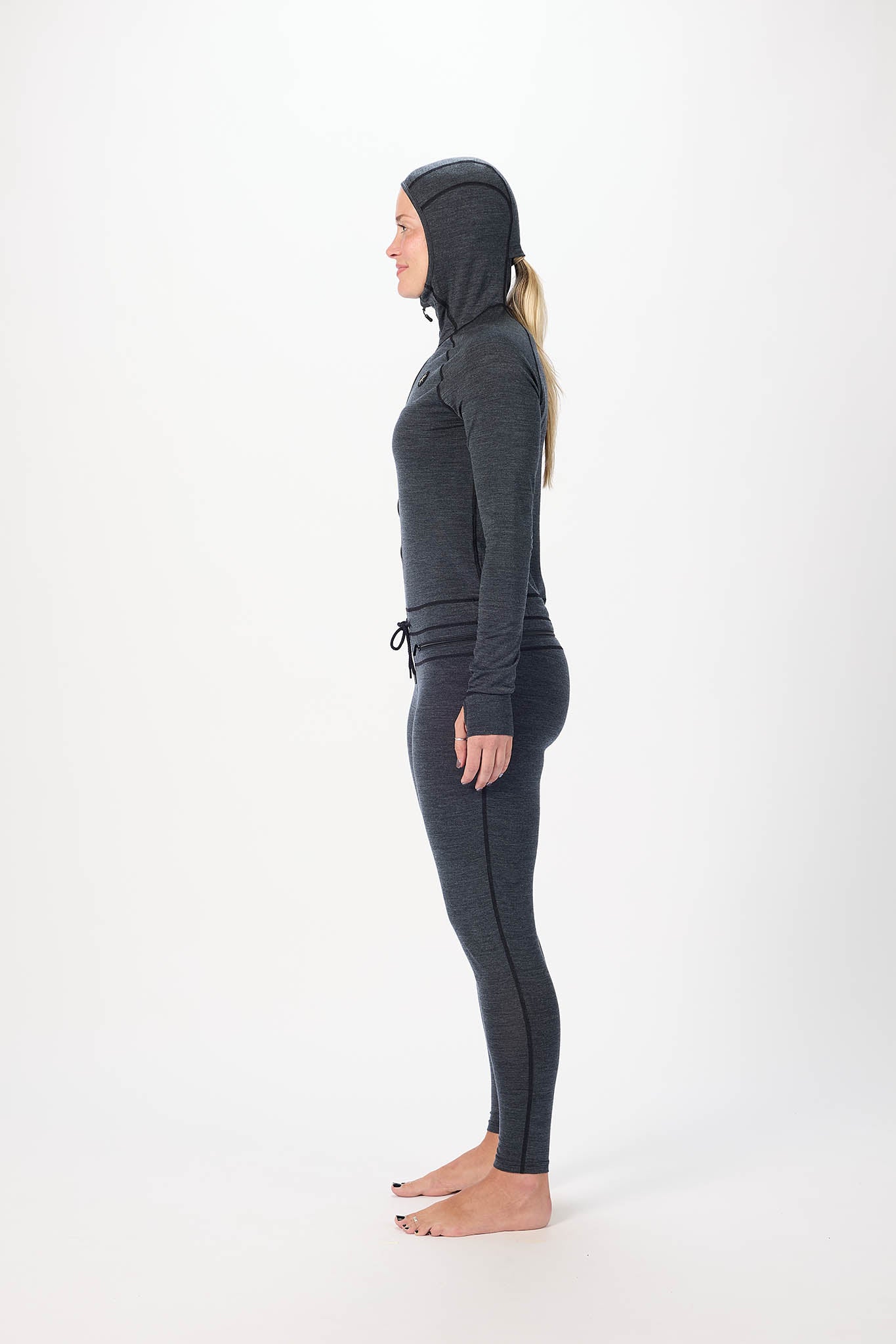 A woman stands in profile wearing a dark gray full-body hooded suit, including a long-sleeve top, leggings, and hood, against a plain white studio background.