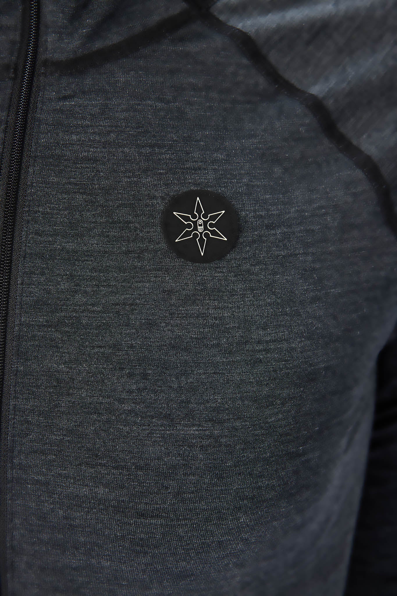 Close-up of a dark heather gray athletic jacket with a black circular chest patch featuring a white compass rose logo.