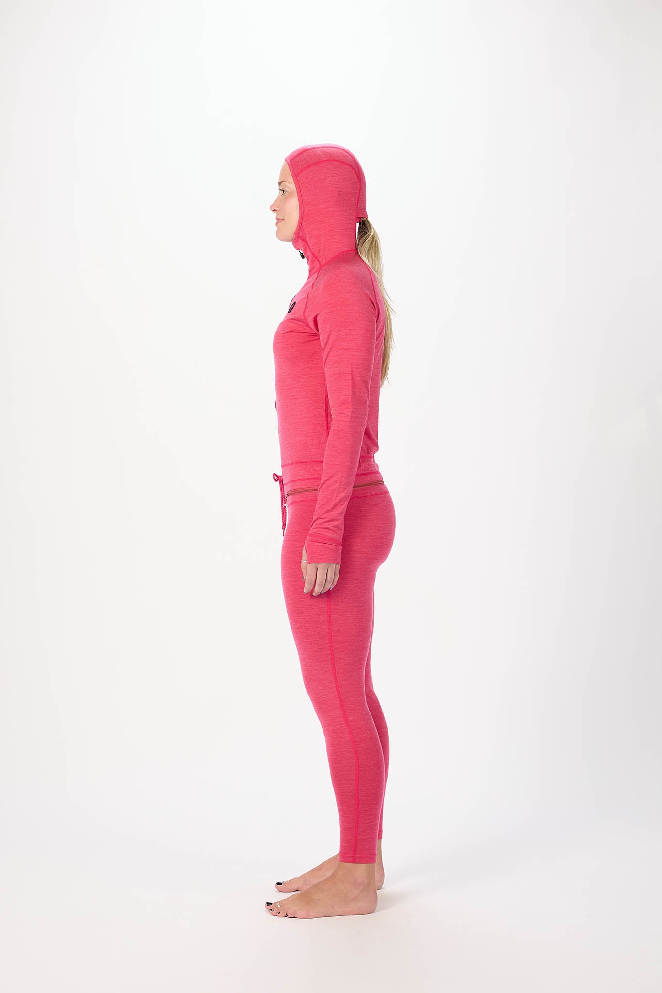 A woman in profile wearing a bright pink full-body hooded one-piece outfit, standing barefoot against a plain white studio background.
