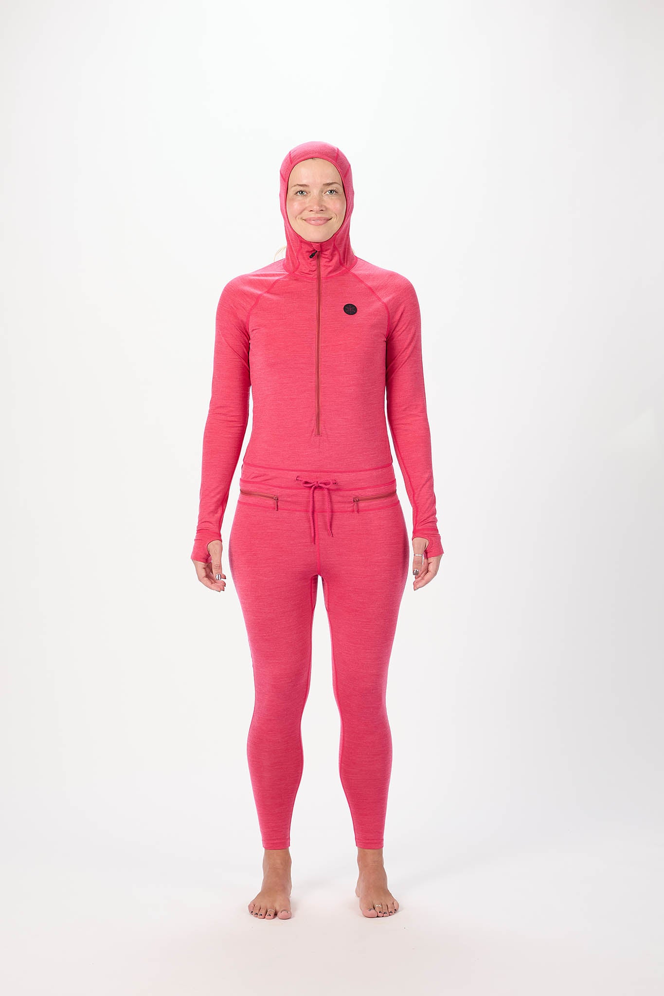 A woman modeling a bright pink full-body hooded suit with a front zipper, long sleeves, and matching leggings against a plain white studio background.