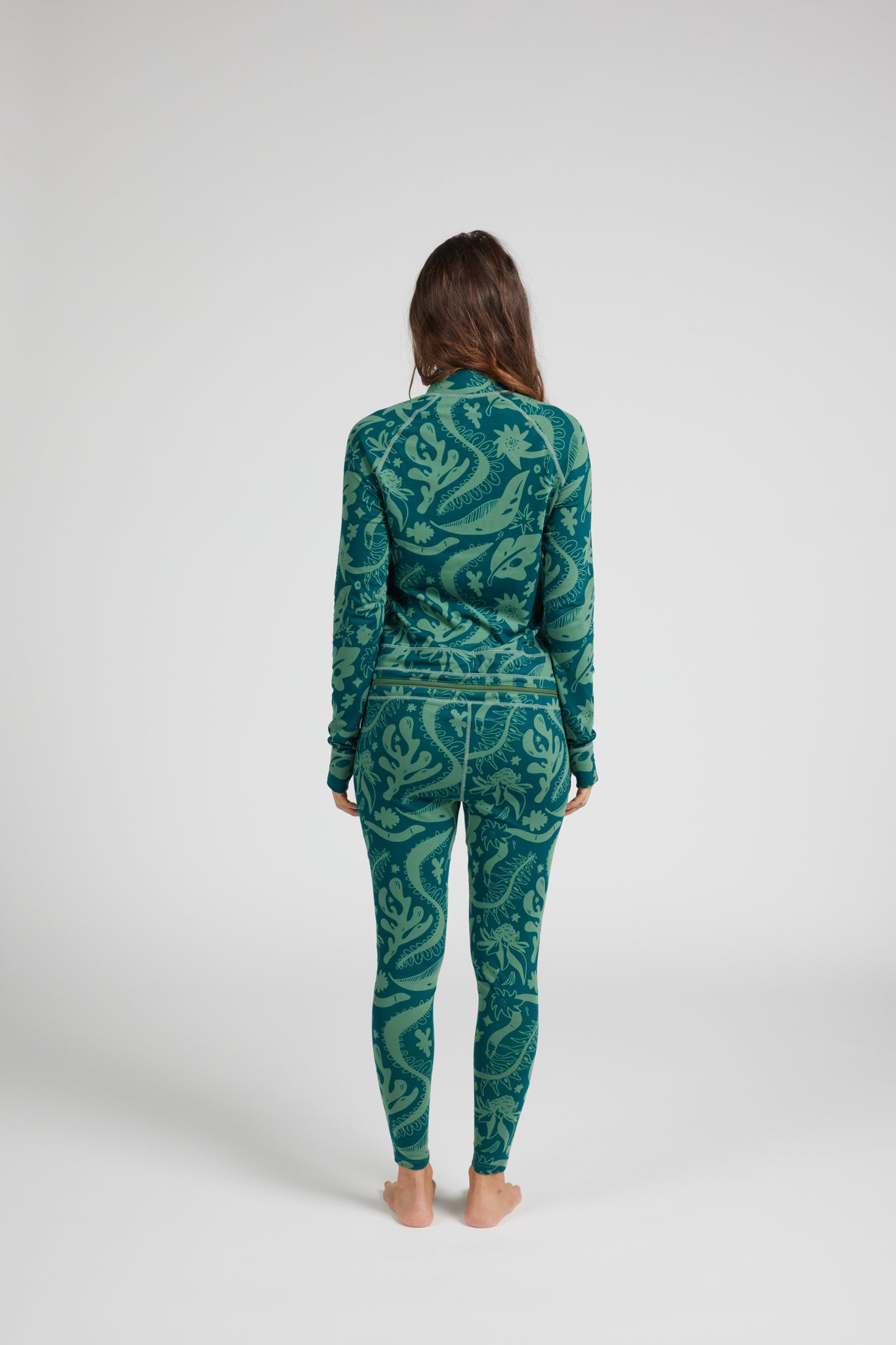A woman stands with her back to the camera wearing a matching long-sleeve top and leggings set in a teal and green paisley pattern, set against a plain light gray studio background.