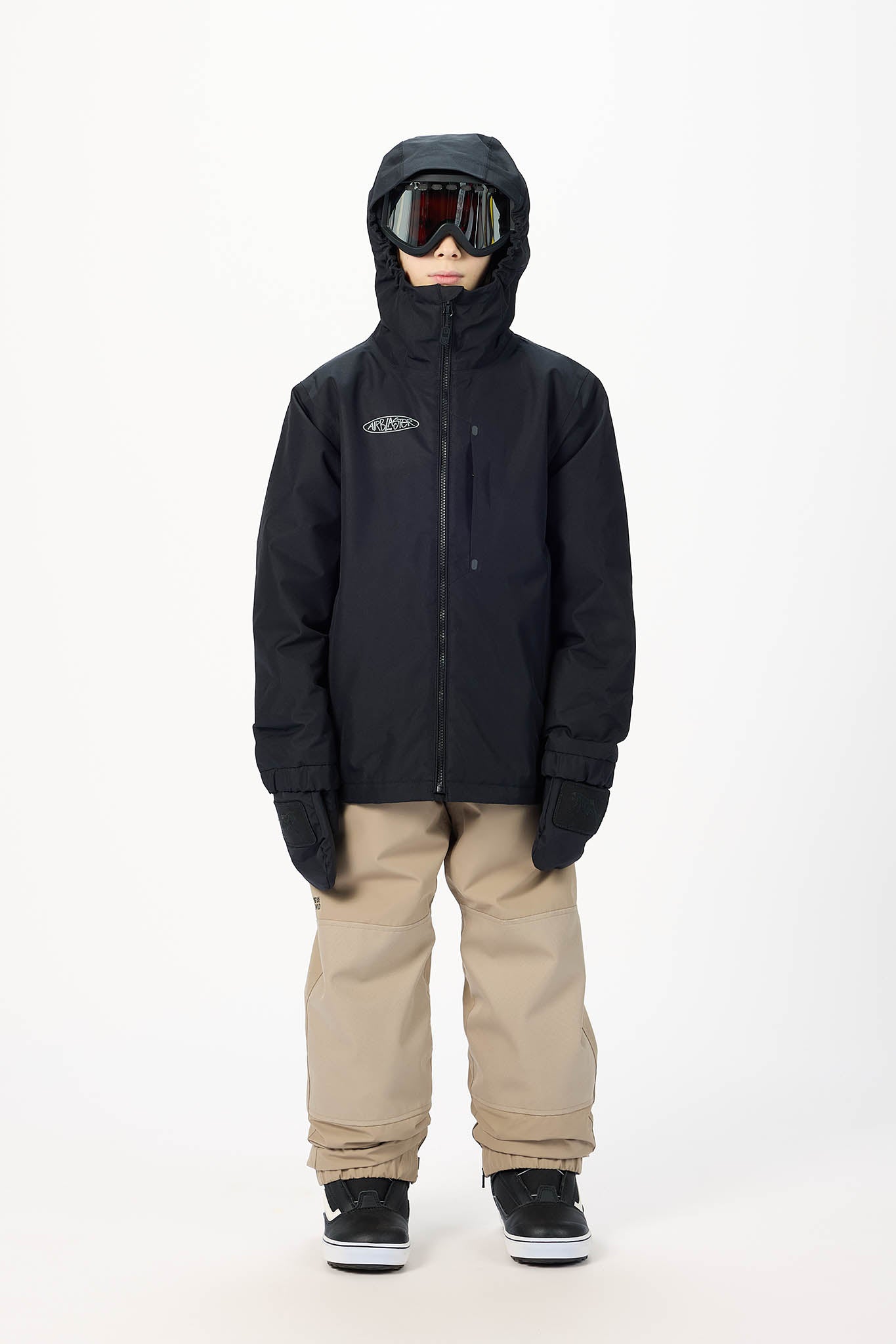A child model wearing a black hooded snowboard jacket, beige snow pants, black and white sneakers, and ski goggles, standing against a plain white studio background.