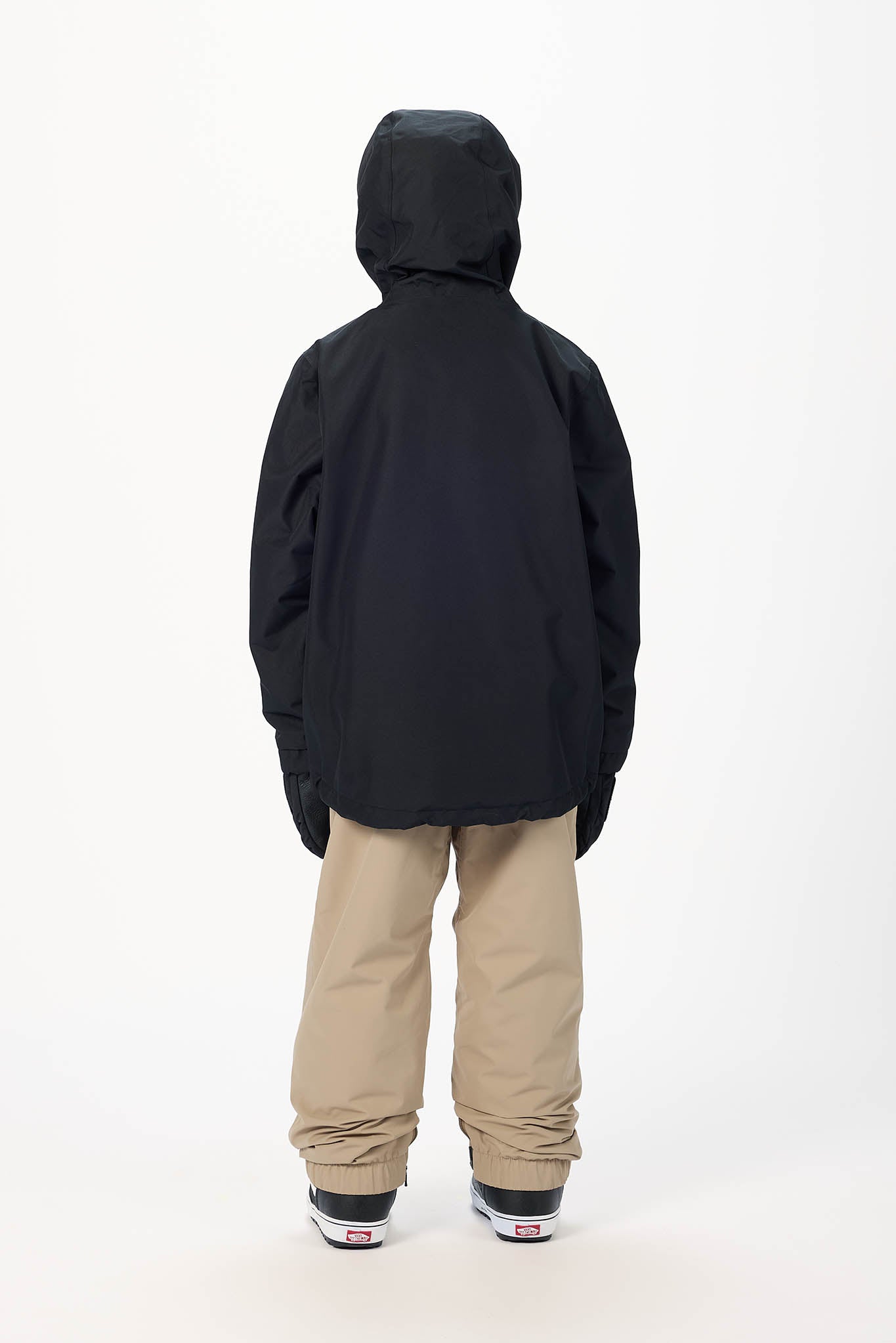 A child viewed from the back wearing a black hooded jacket, beige snow pants, and black sneakers with white soles, standing against a plain white studio background.