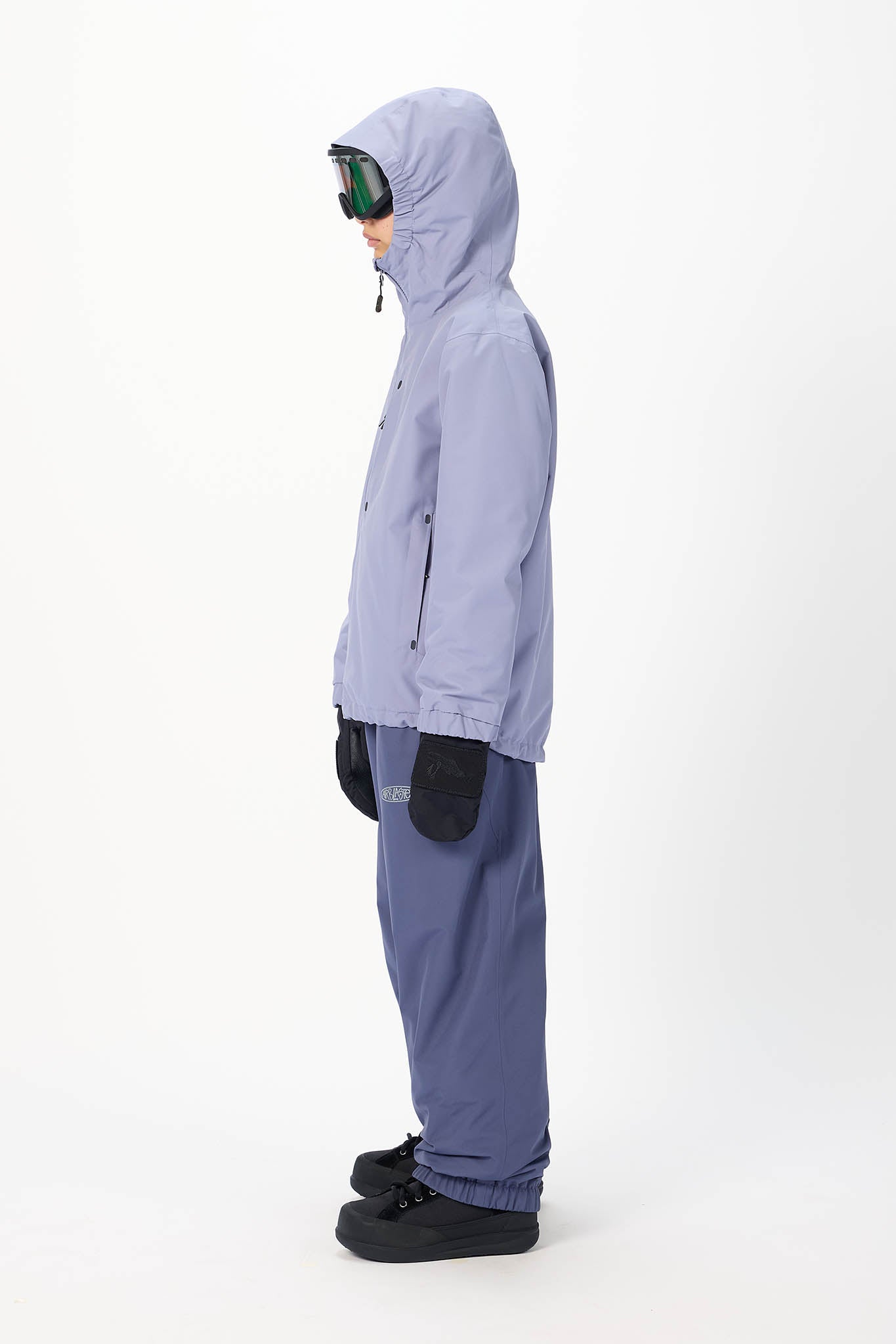 A child model shown in profile wearing a light periwinkle hooded jacket, matching pants, black gloves, and black snow boots, with ski goggles resting on the hood, against a plain white studio background.