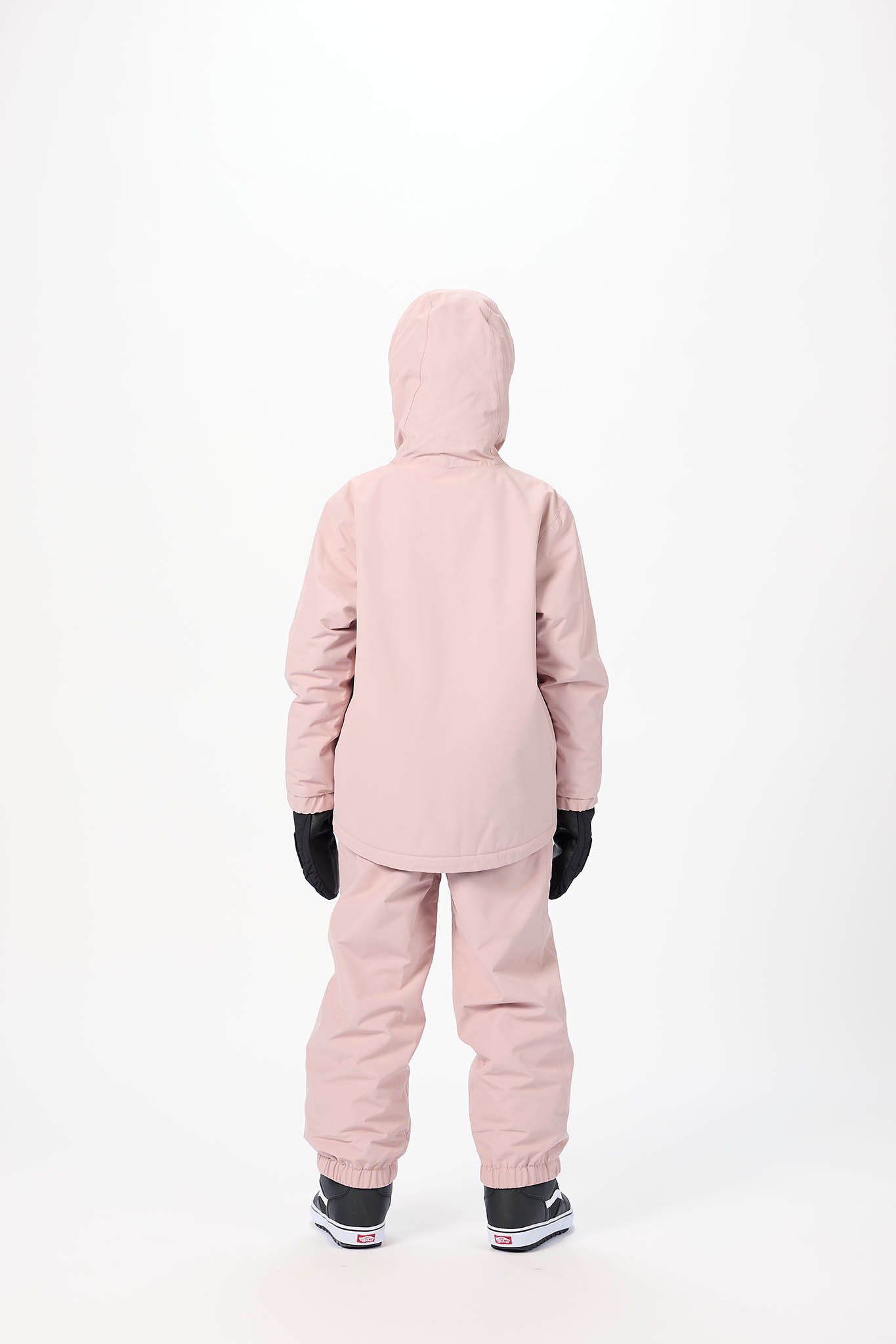 A child viewed from the back wearing a light dusty pink hooded snowsuit with black gloves and black-and-white sneakers, standing against a plain white studio background.