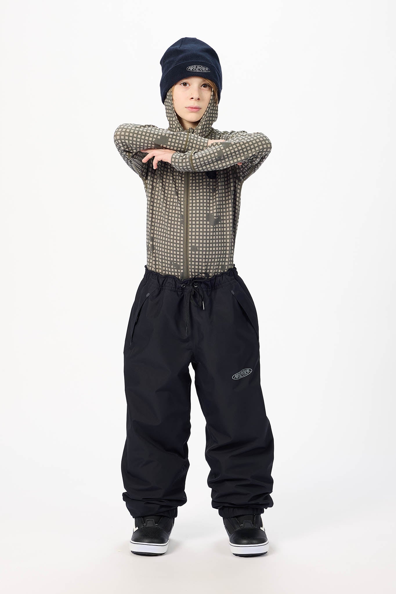 A child model wearing a dark navy beanie, a brown and gray patterned zip-up hoodie, black snow pants, and black sneakers, posing with arms crossed against a plain white studio background.