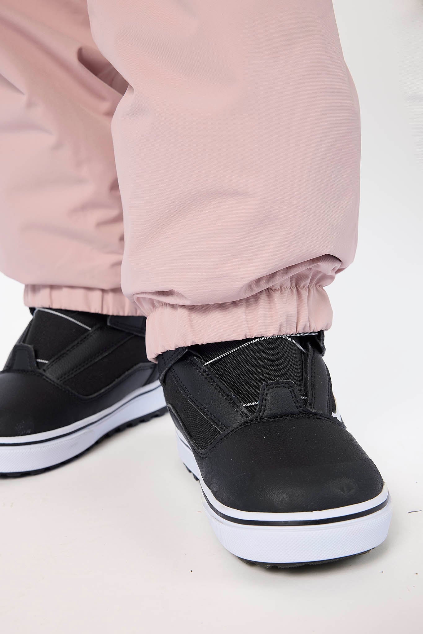 A close-up of a child's lower legs wearing dusty pink snow pants and black winter boots with white soles, standing against a plain white background.