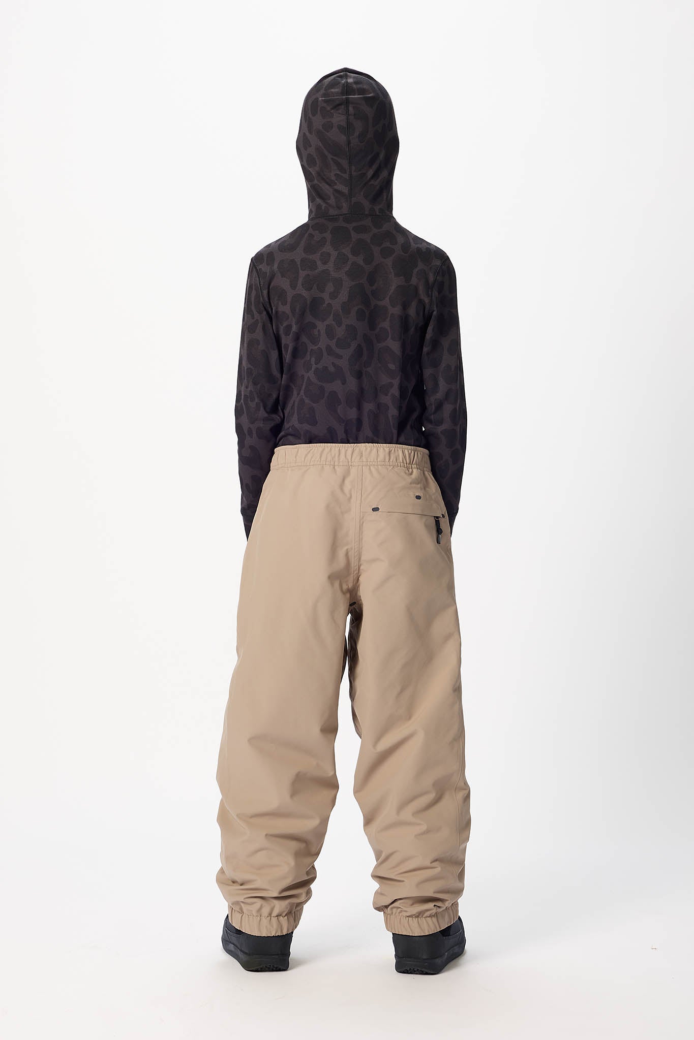 A child viewed from the back wearing a black hooded sweatshirt with a subtle tonal animal pattern and beige snow pants, standing against a plain white studio background.