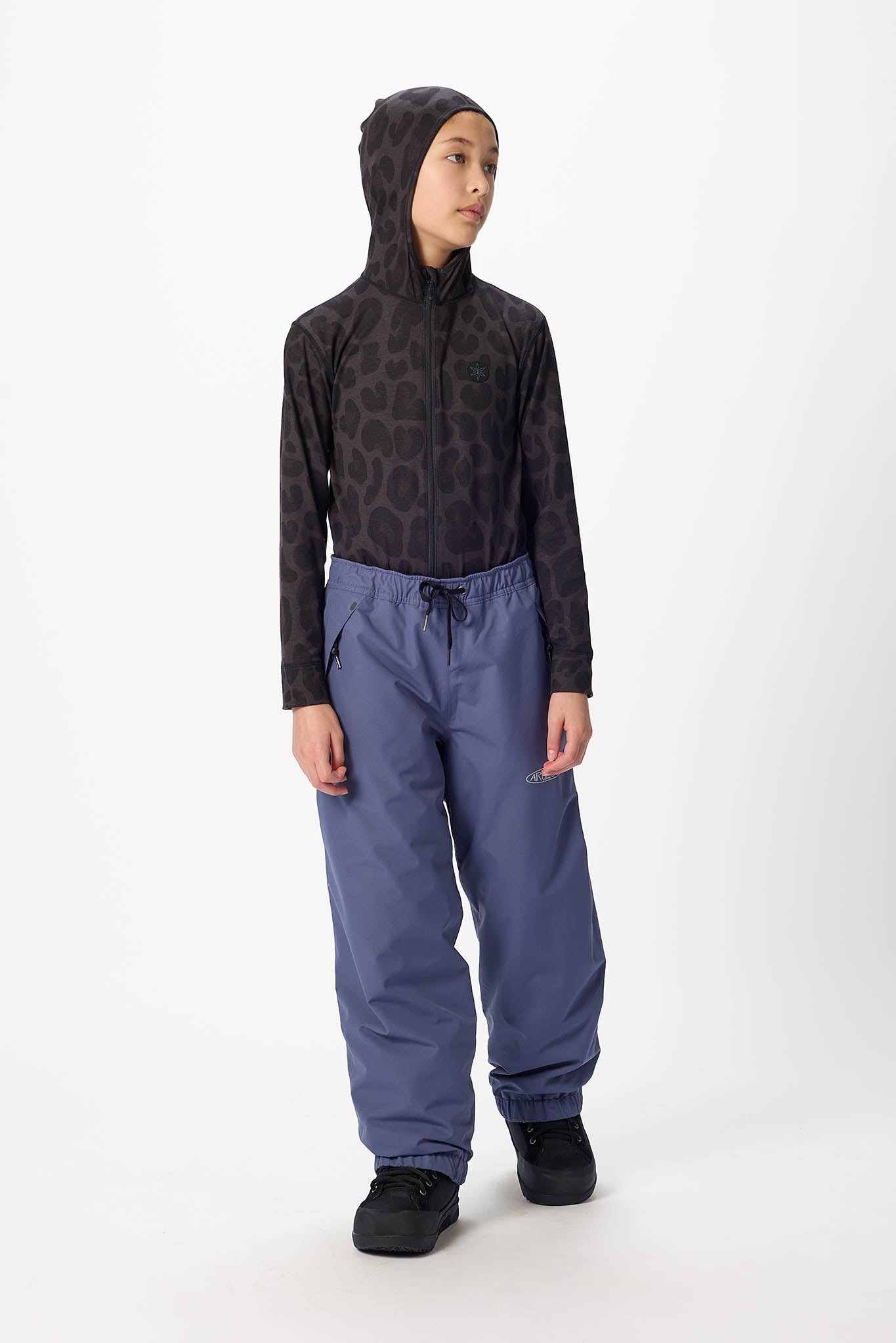 A child model wearing a black hooded sweatshirt with a subtle tonal pattern on the upper body and sleeves, paired with slate blue pants and black shoes, standing against a plain white studio background.