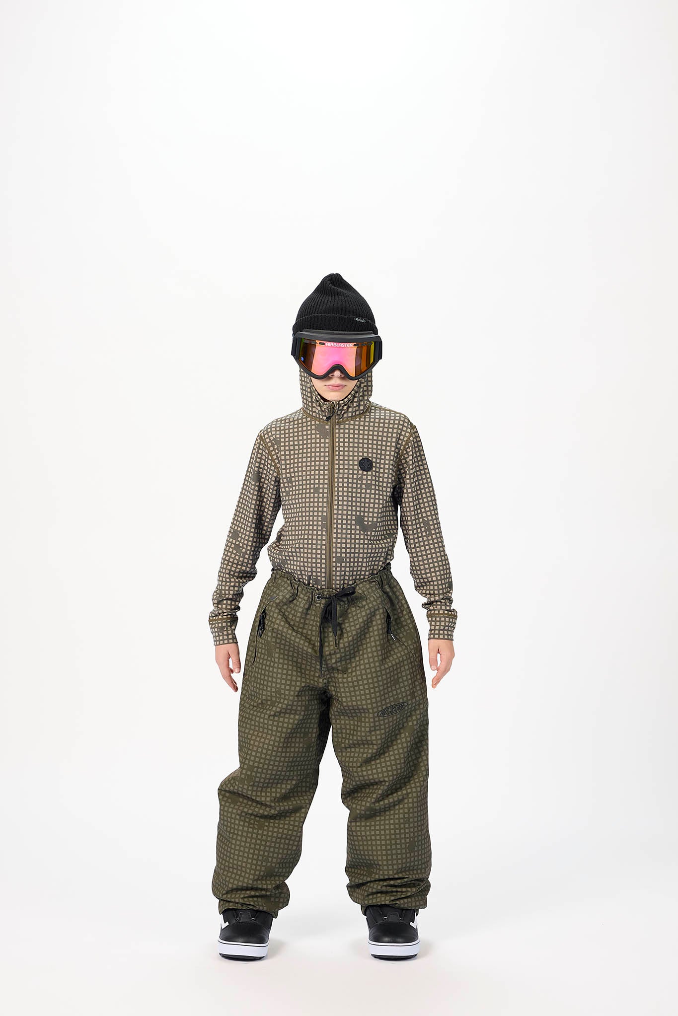 A child model wearing a matching olive green and black checkered snowboard jacket and bib pants, styled with a black beanie, pink-tinted goggles, and black sneakers, standing against a plain white studio background.