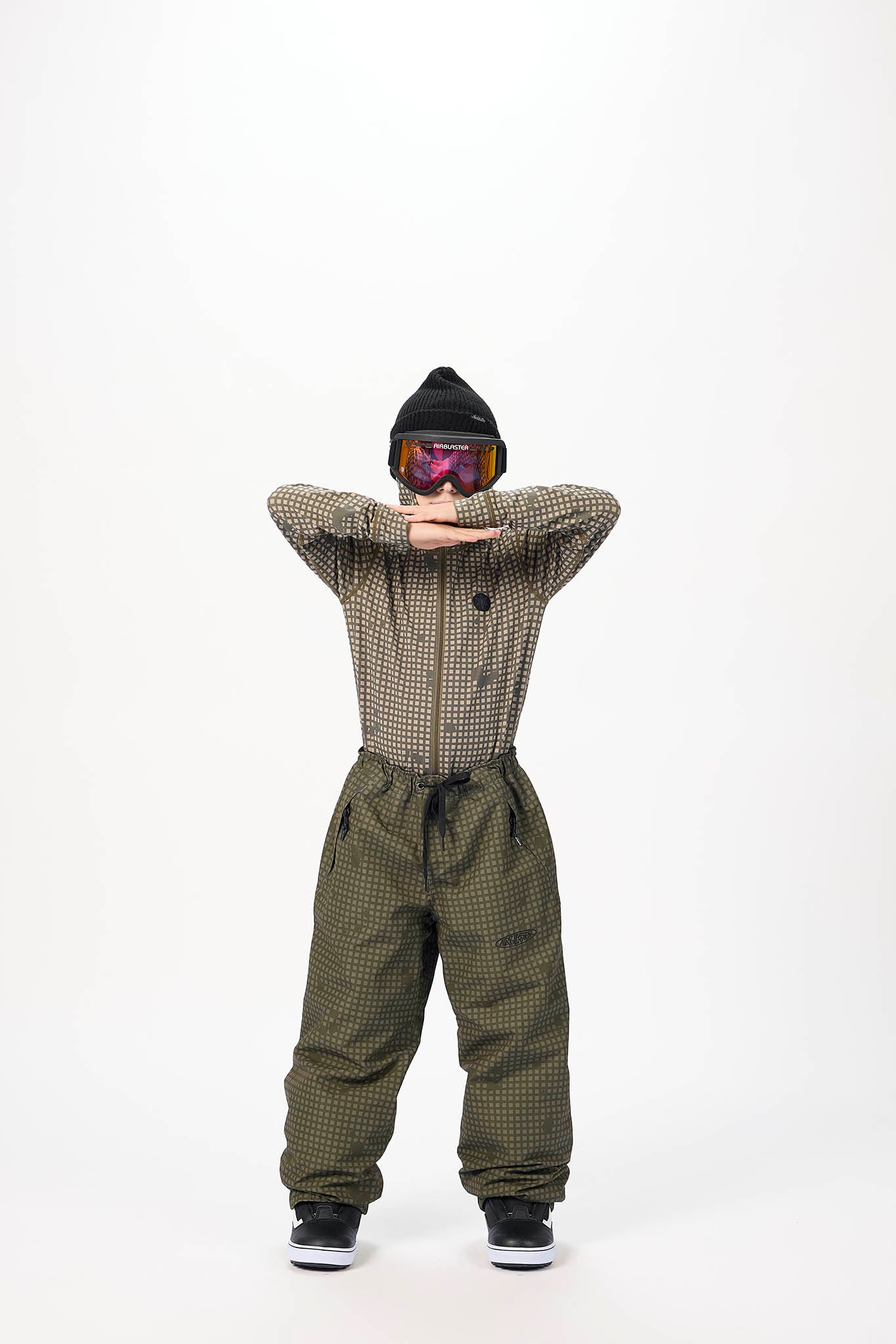A child wearing a black beanie, ski goggles, a brown and black checkered zip-up jacket, and olive green checkered snow pants, posing against a solid white background.
