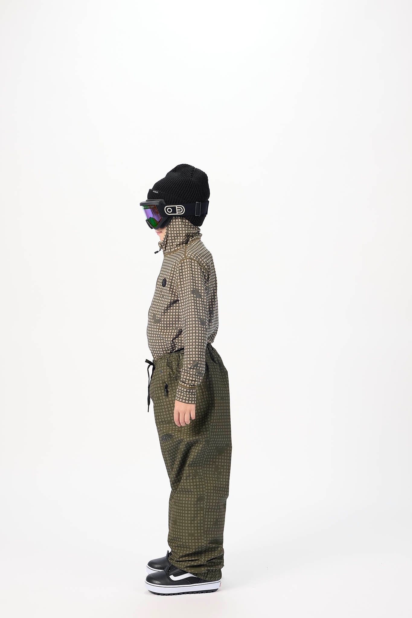 A child standing in profile wearing a black beanie, ski goggles, a brown and black checkered long-sleeve top, olive green checkered snow pants, and black sneakers against a plain white studio background.