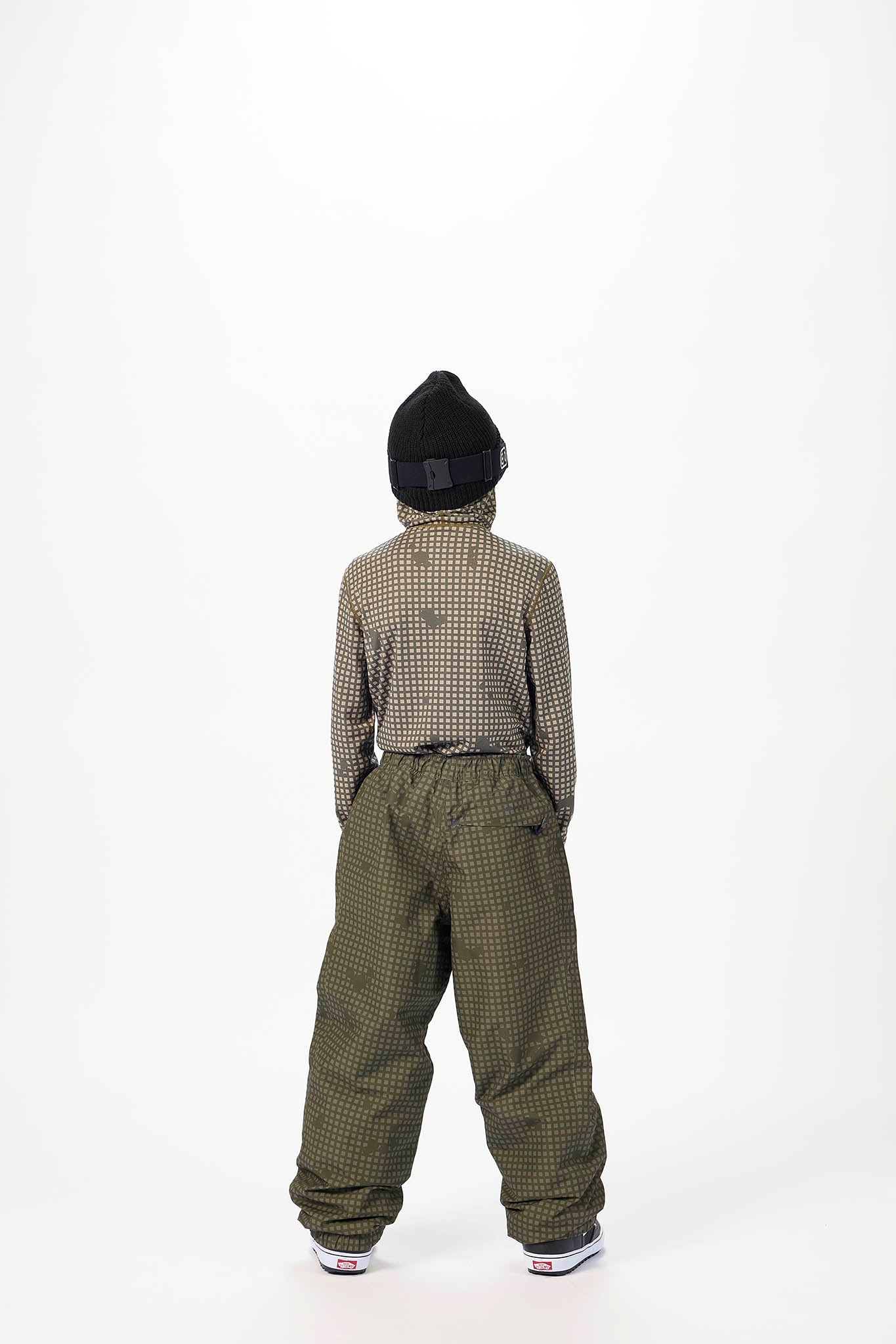 A child shown from the back wearing a black knit beanie, a brown and black checkered long-sleeve top, and olive green checkered snow pants, standing against a plain white studio background.