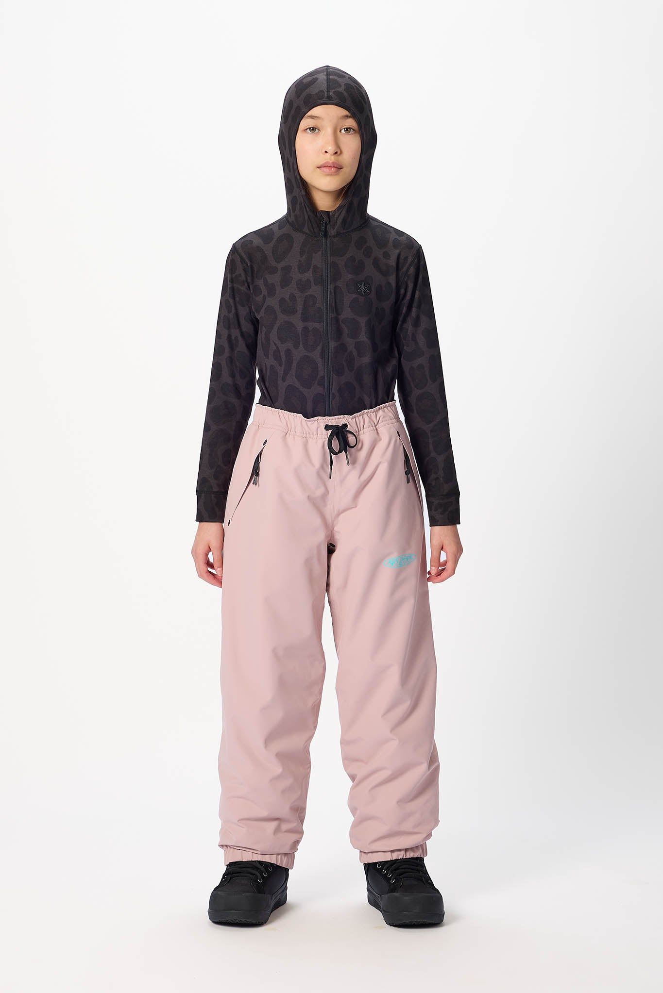 A child model stands against a light gray studio background wearing a dark patterned hooded long-sleeve top and light pink snow pants with black shoes.