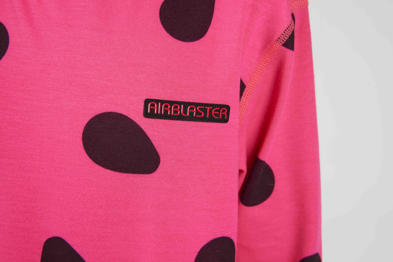 A close-up of a bright pink long-sleeve shirt with large dark brown spots and a black rectangular patch on the chest that reads 'AIRBLASTER' in red.