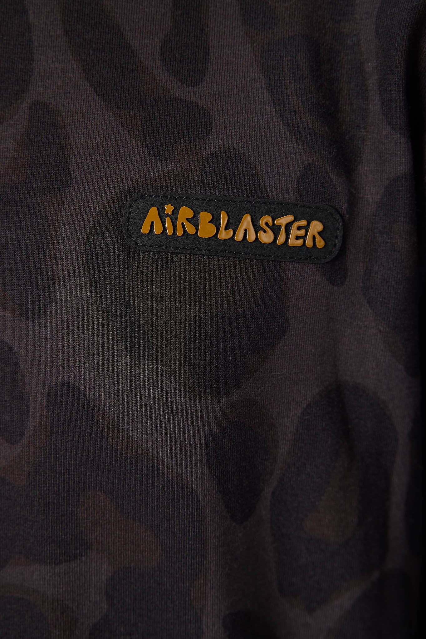 Close-up of a dark camouflage-patterned Airblaster sweatshirt featuring a black patch with the brand name 'AIRBLASTER' in orange-yellow lettering.