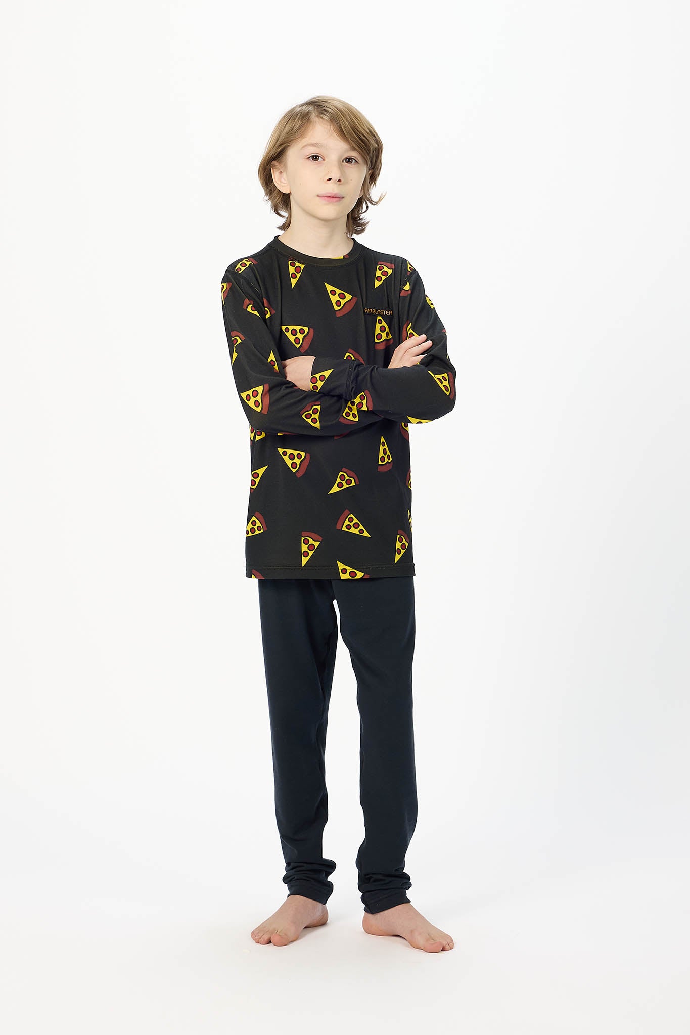 A young boy stands with arms crossed wearing a black long-sleeve pajama top with a yellow and red pizza slice pattern, paired with black pants, against a plain white studio background.