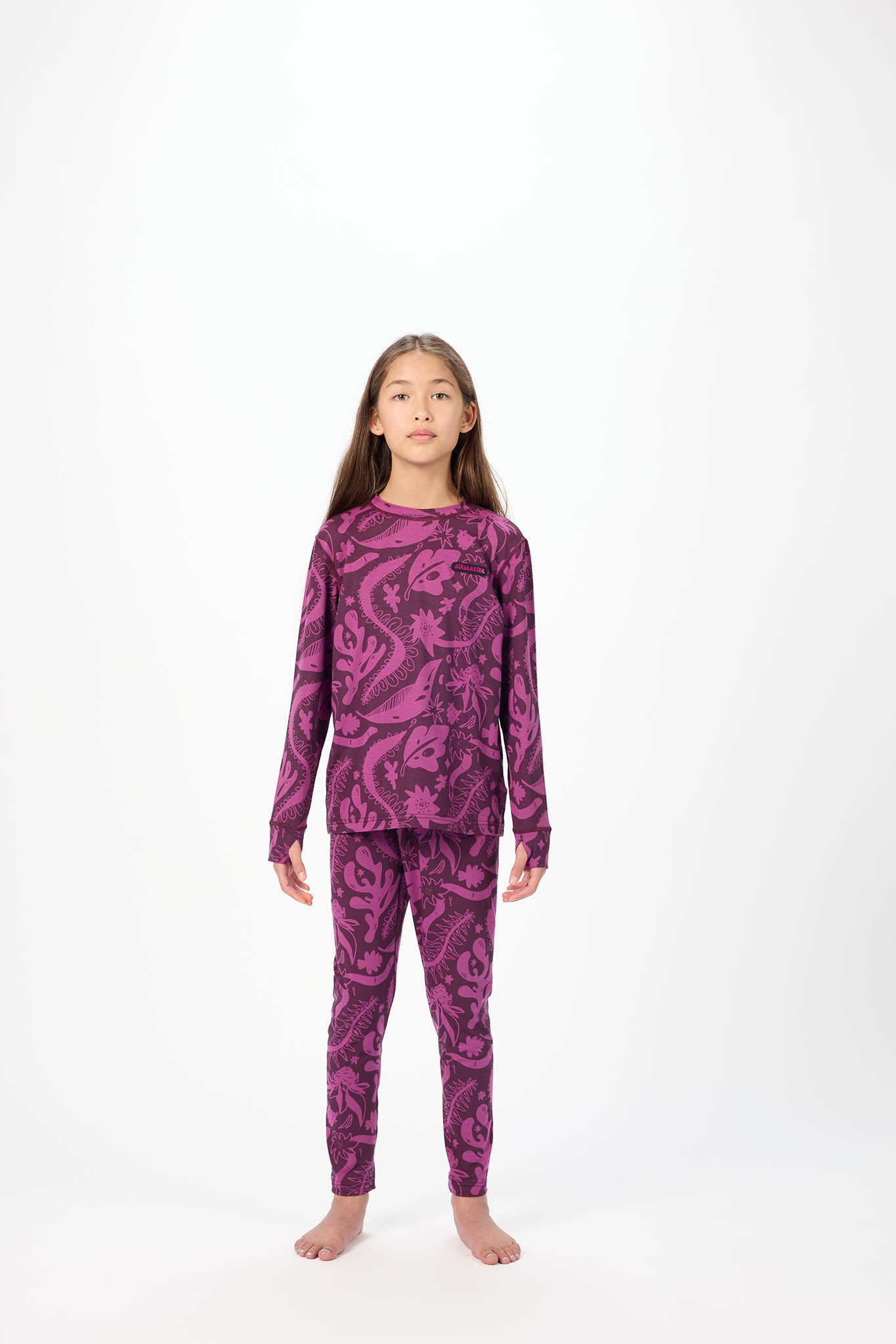 A young girl stands barefoot against a plain white studio background, wearing a matching long-sleeve top and pants set in a dark purple paisley pattern.