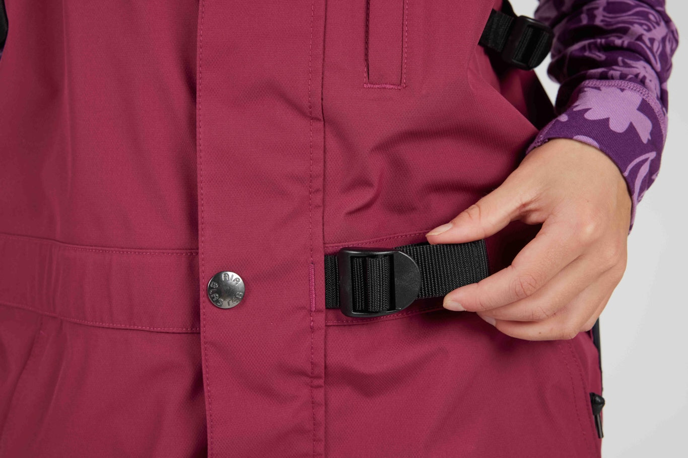 Close-up of a person wearing a burgundy insulated jacket and purple patterned long-sleeve shirt, adjusting a black webbing belt on the jacket.