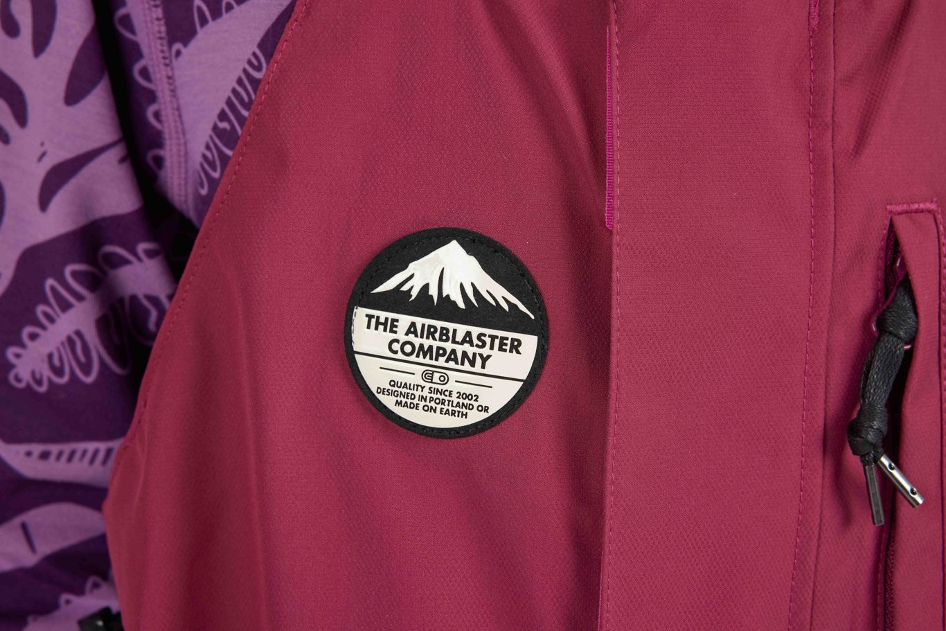 A close-up of a dusty rose pink jacket featuring a circular black and white 'The Airblaster Company' patch on the chest, with a purple patterned garment partially visible on the left.