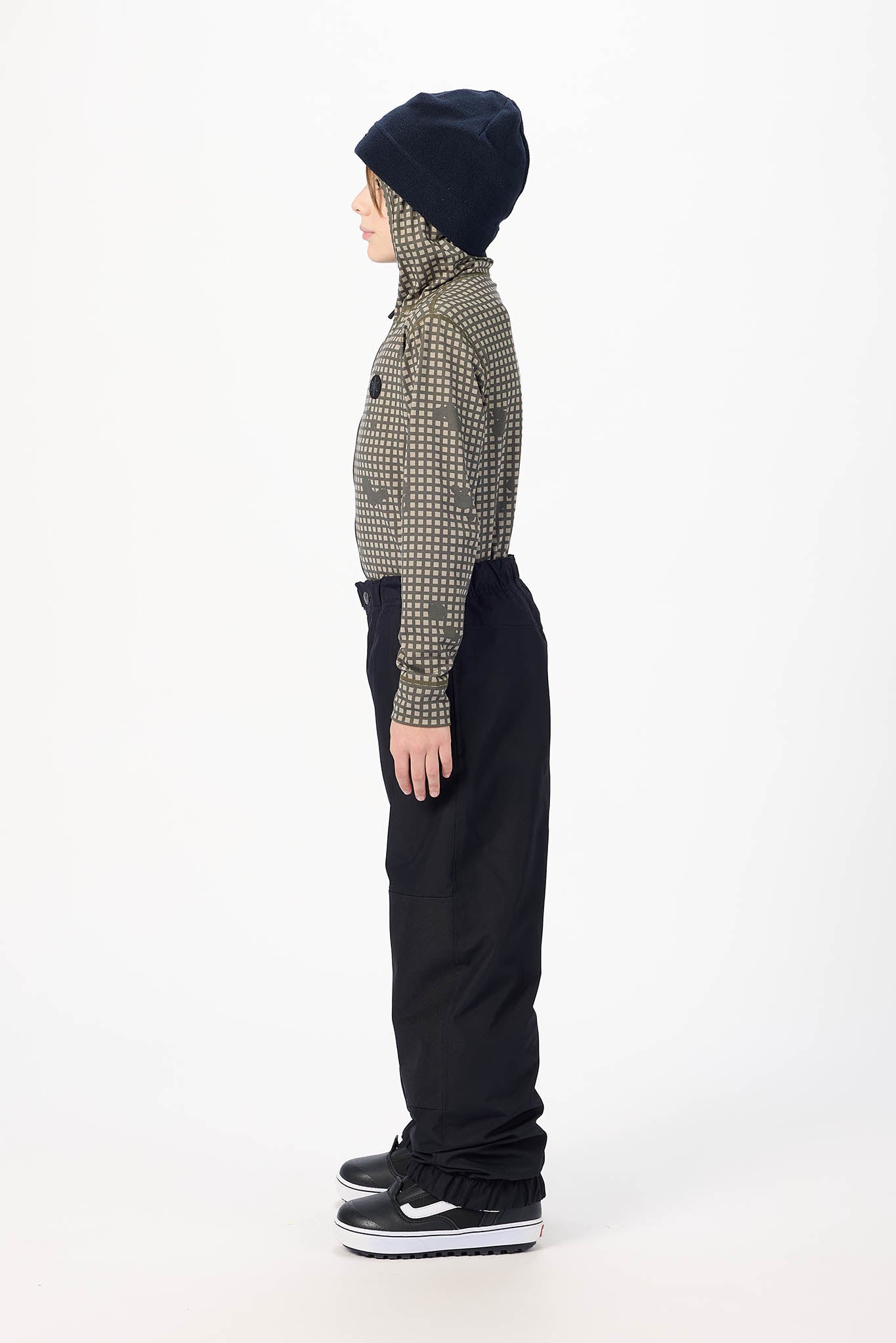 A child model shown in profile wearing a dark navy beanie, a patterned long-sleeve top, black pants, and black sneakers with white soles, standing against a plain white studio background.