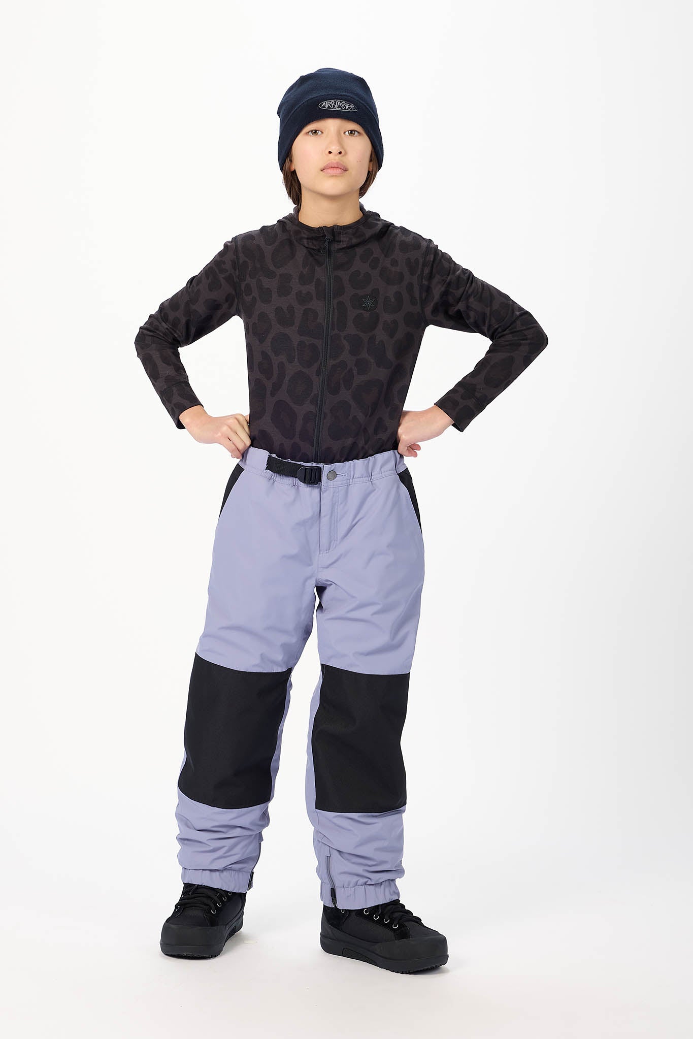 A child model wearing a black patterned long-sleeve top, light purple and black snow pants, a dark beanie, and black snow boots, standing against a white studio background.
