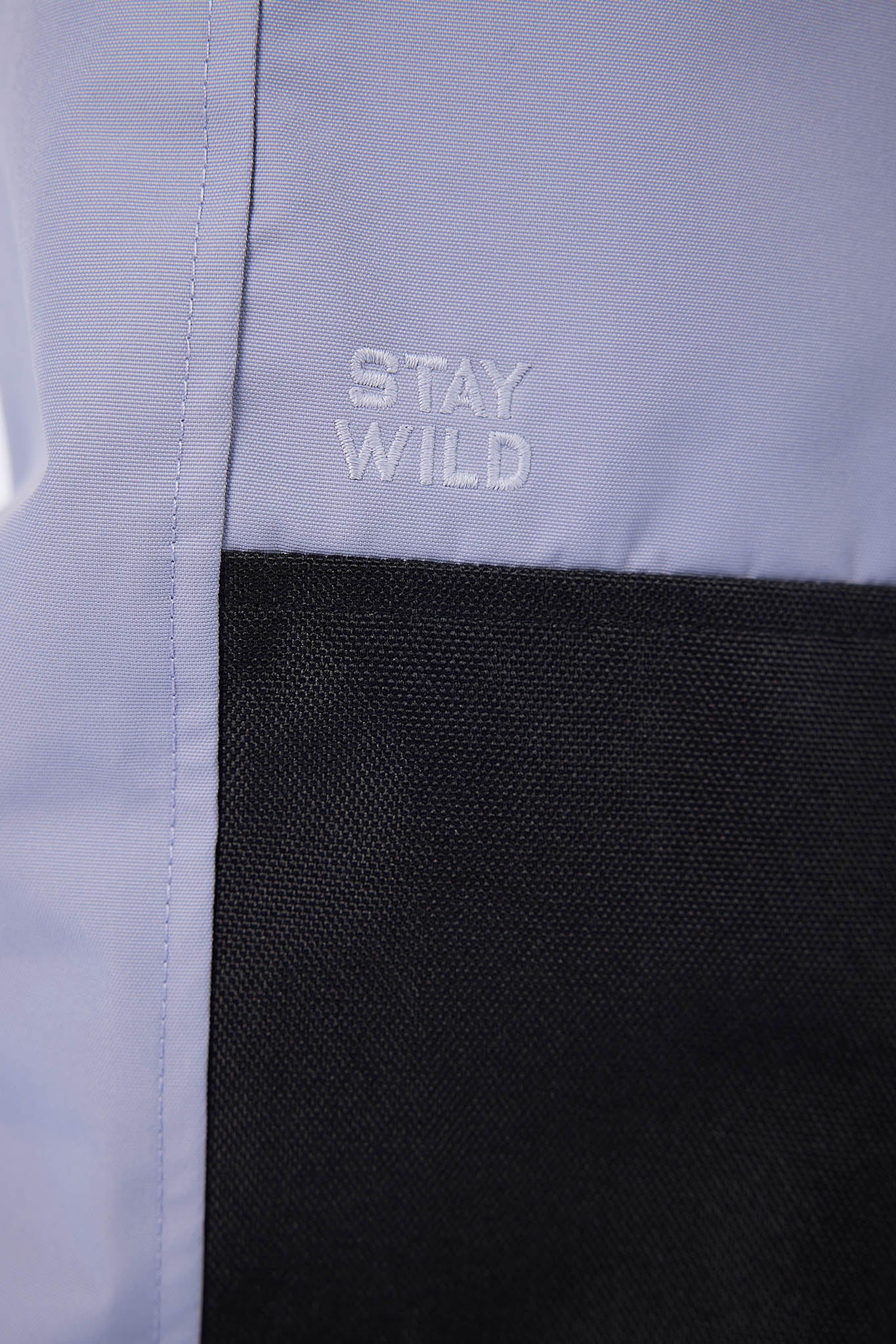Close-up of a color-blocked jacket or fabric panel with a light lavender-gray upper section and a black lower mesh panel. The words 'STAY WILD' are embroidered in light gray on the upper section.