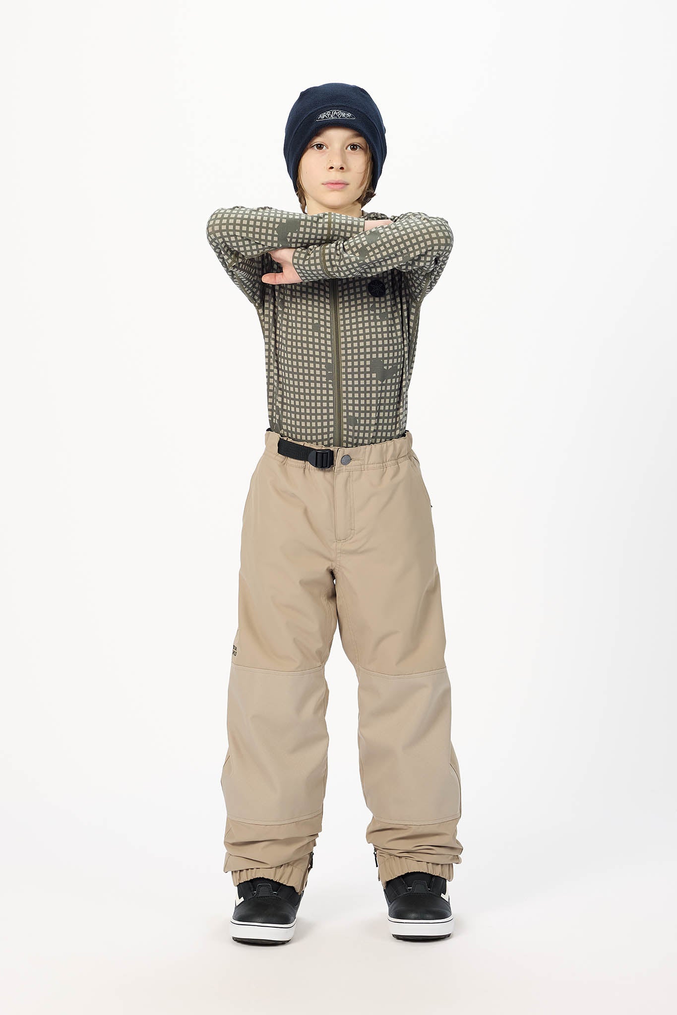 A child model wearing beige snow pants, a green and black patterned long-sleeve shirt, a black beanie, and black sneakers, standing against a plain white studio background.