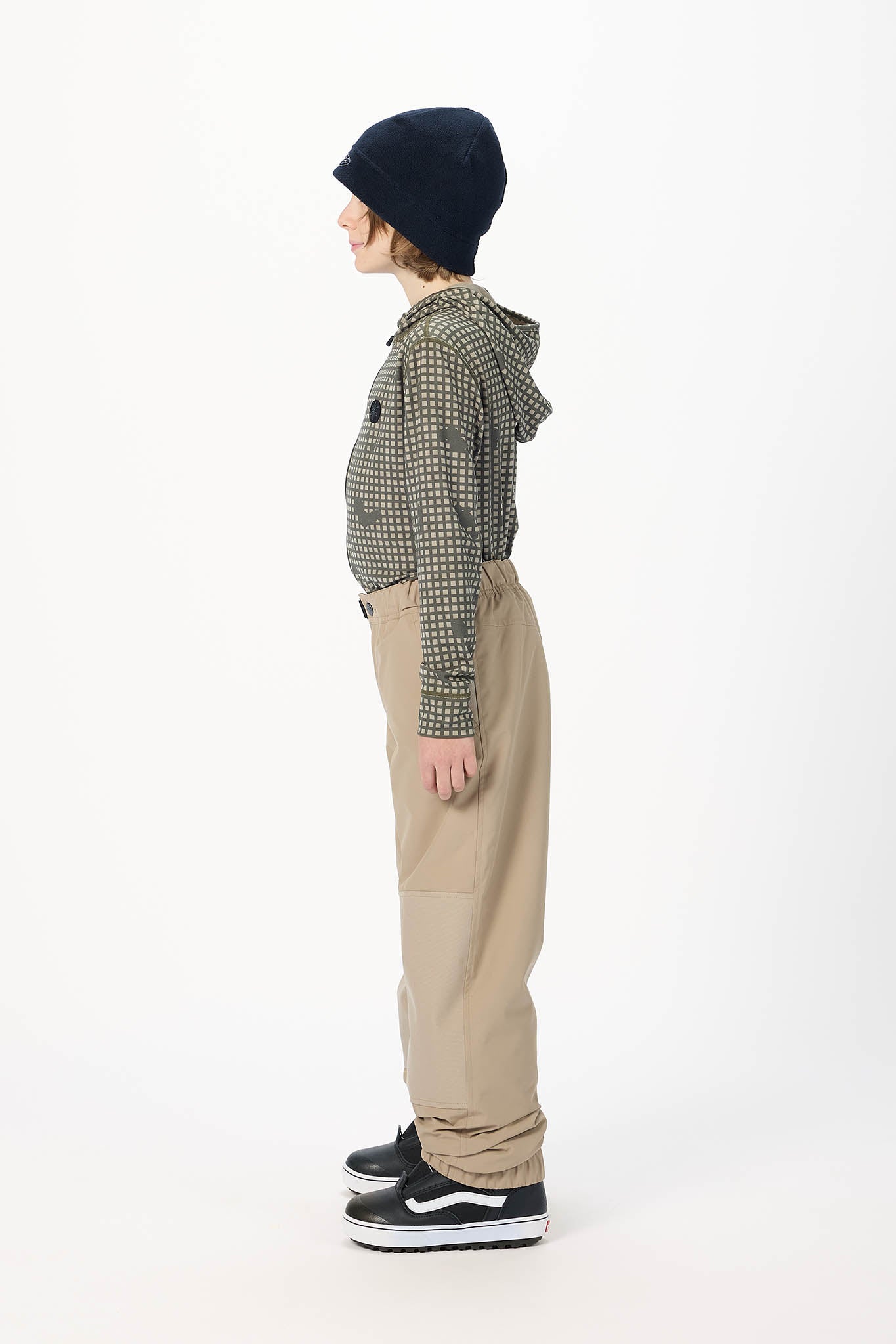 A child model shown in profile wearing a black knit beanie, a green and black checkered long-sleeve top, beige cargo-style pants, and black sneakers with white soles, against a plain white studio background.