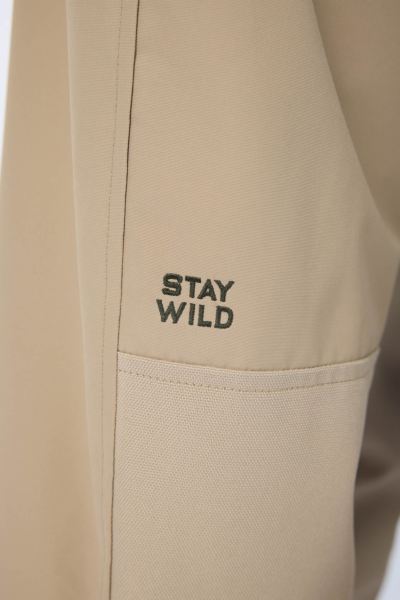 Close-up of a beige jacket or pants sleeve with a pocket and the words 'STAY WILD' printed in black on the fabric.