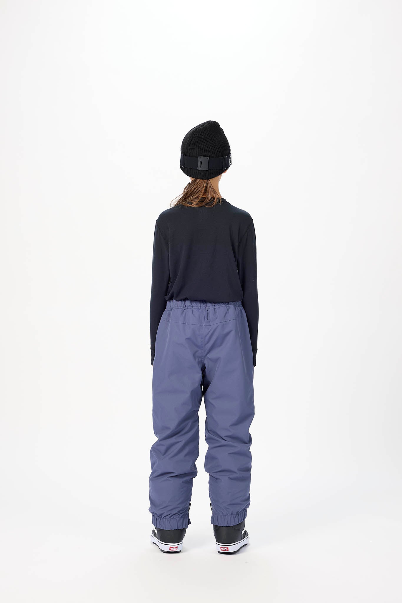 A child viewed from behind wearing a black beanie, a black long-sleeve top, and slate blue snow pants against a plain white studio background.