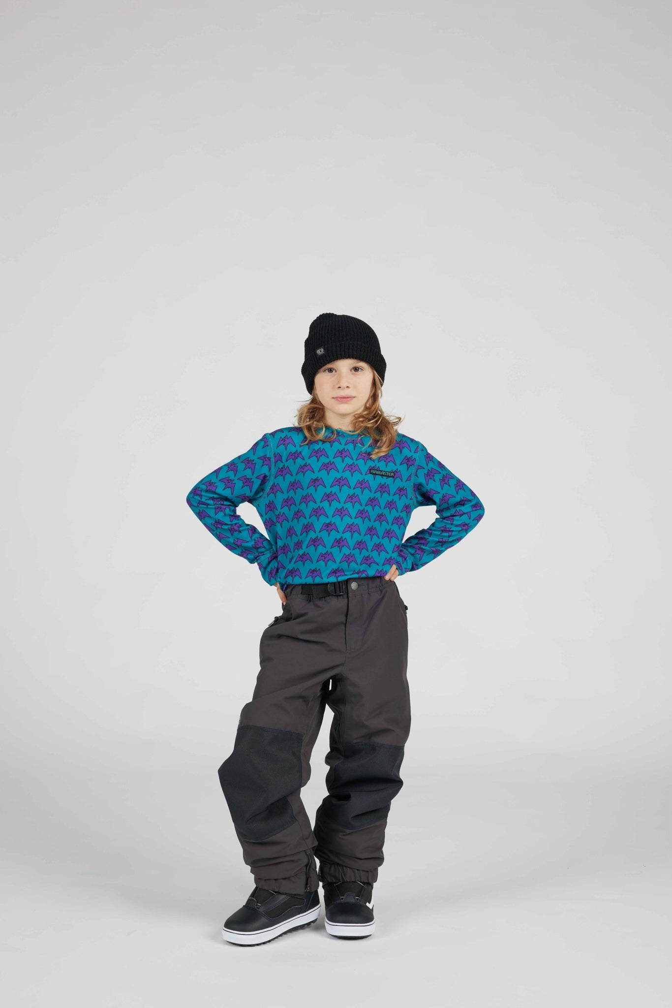 A child model wearing a blue and black patterned long-sleeve top, dark gray snow pants, a black beanie, and black sneakers, standing against a light gray studio background.