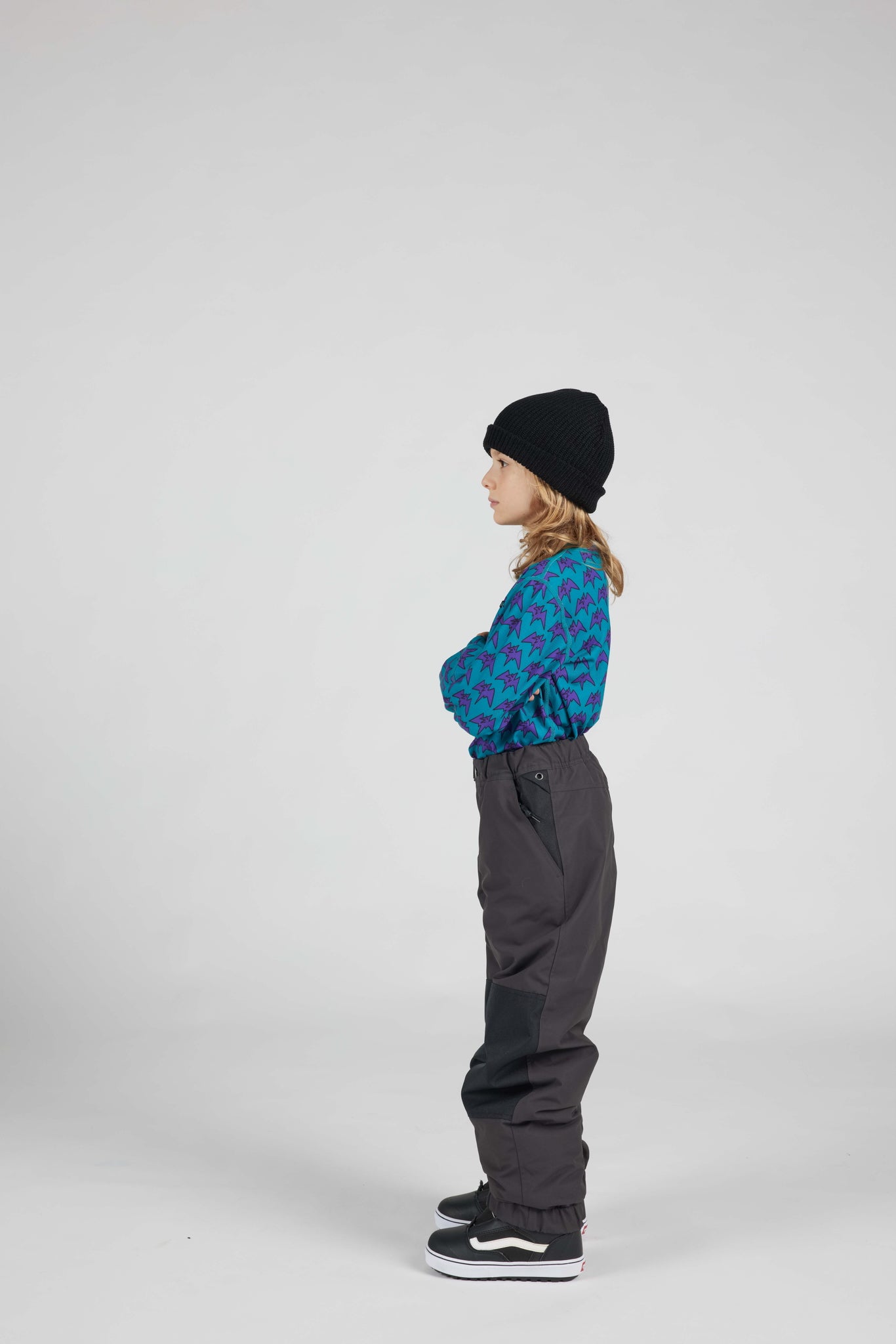 A child standing in profile against a light gray studio background, wearing a black beanie, a blue and red patterned long-sleeve top, dark gray pants, and black sneakers.