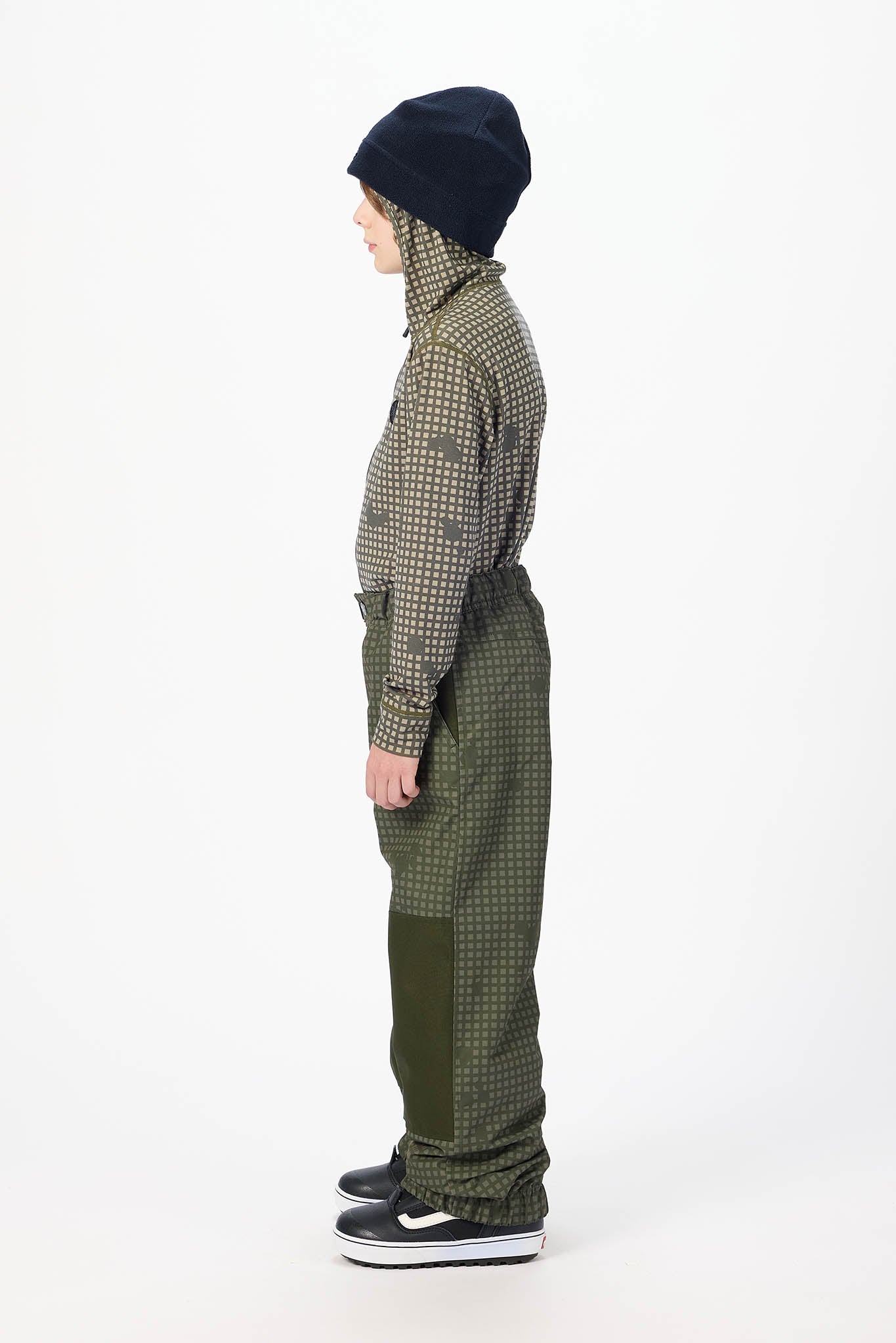 A child shown in profile wearing a dark navy beanie, a green and beige patterned jacket, olive green bib-style pants, and black sneakers against a plain white studio background.