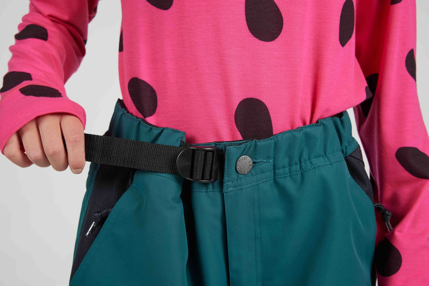 Close-up of a person wearing teal snow pants with a black belt and buckle, paired with a bright pink long-sleeve top with dark spots.