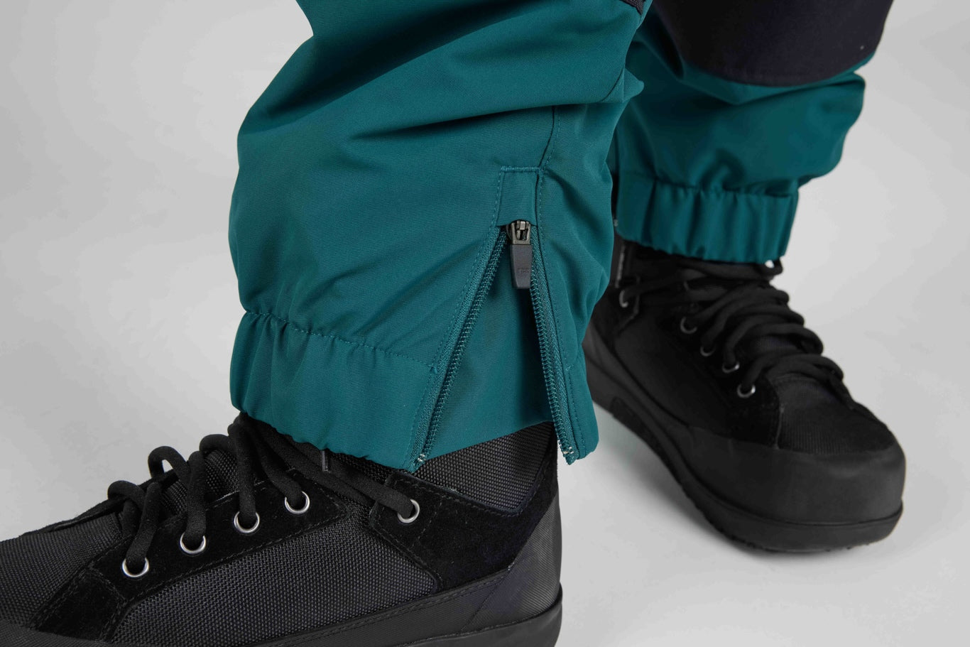 Close-up of teal pants cuffed above black lace-up boots, showing a zippered ankle detail and elastic cuffs on a light gray studio background.