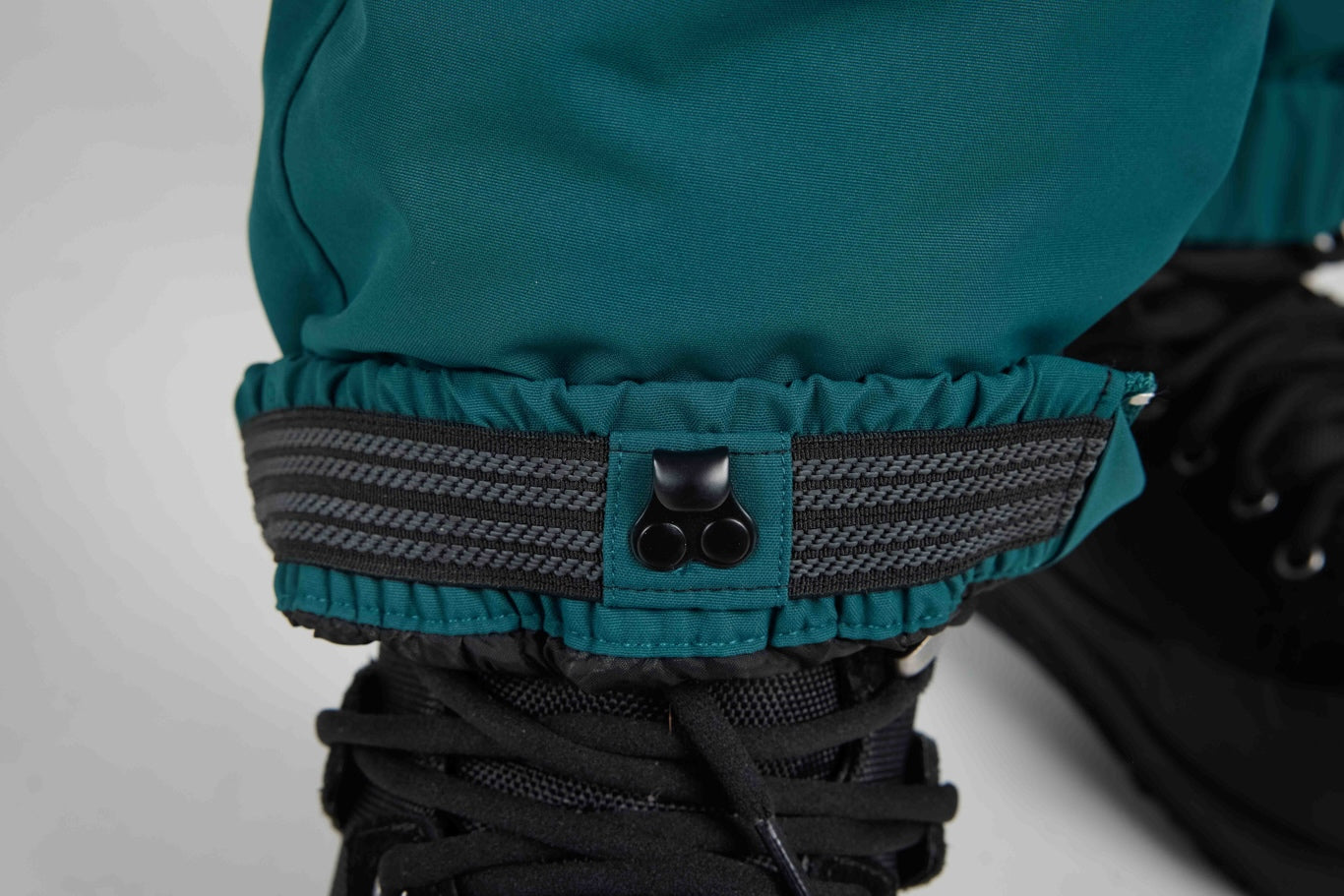 Close-up of a teal waterproof pant cuff with a black elastic gaiter strap and hook-and-loop closure, shown over a black boot.