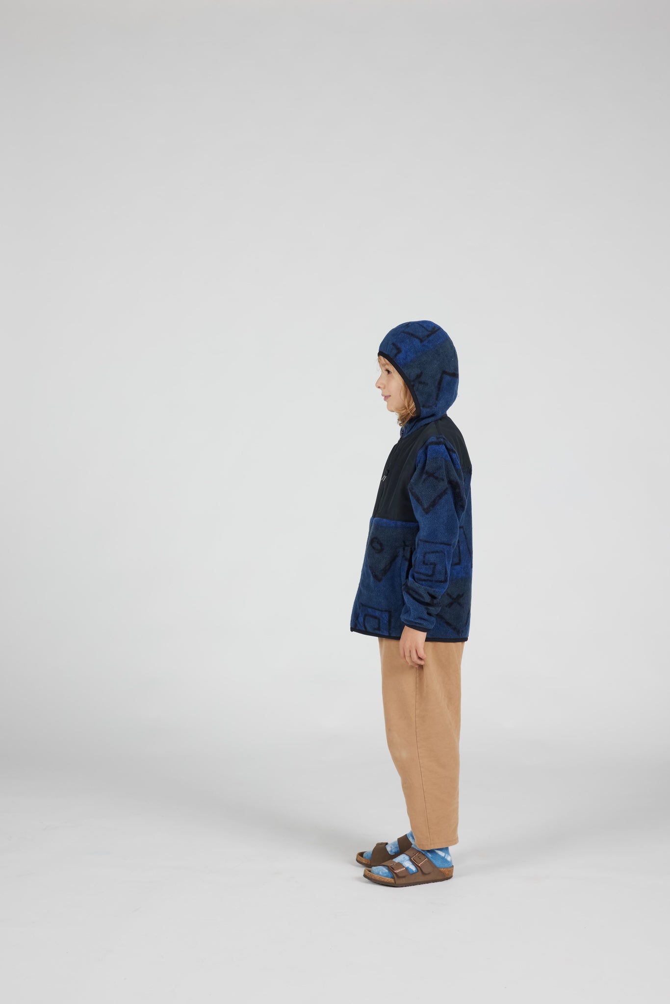 A child standing in profile wearing a dark blue hooded jacket with a black chest panel, tan pants, and sandals, photographed against a plain light gray studio background.