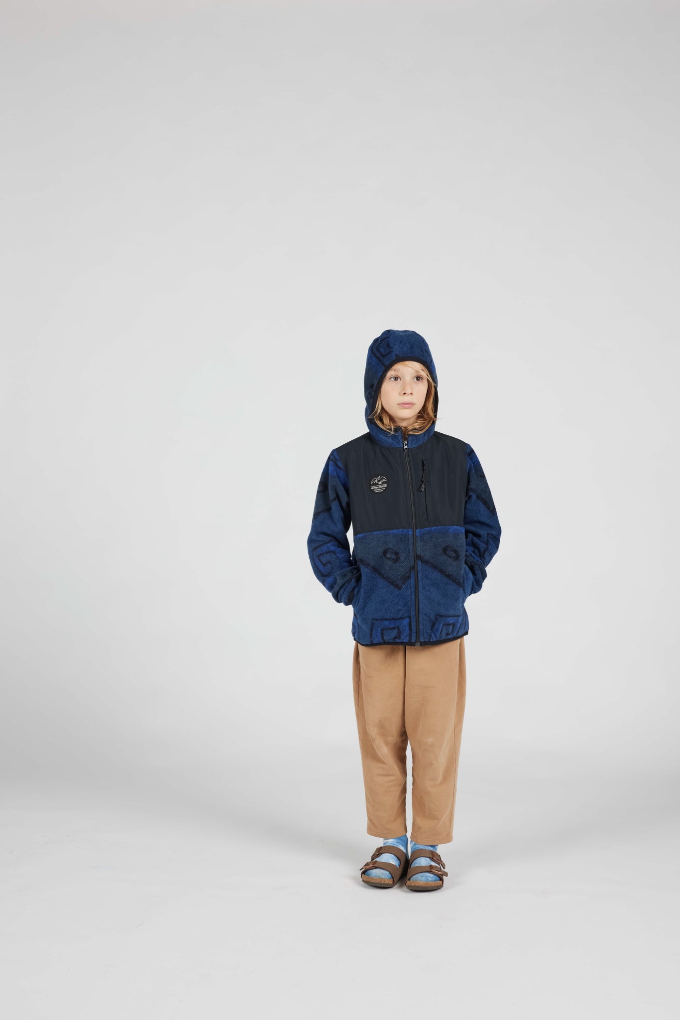 A child model wearing a two-tone navy and blue hooded jacket, tan pants, and sandals, standing against a seamless light gray studio background.