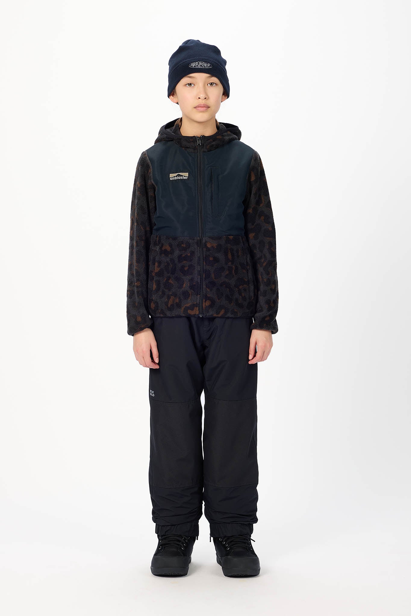 A child model wearing a dark navy and black patterned hooded jacket, black snow pants, and a dark beanie, standing against a plain white studio background.
